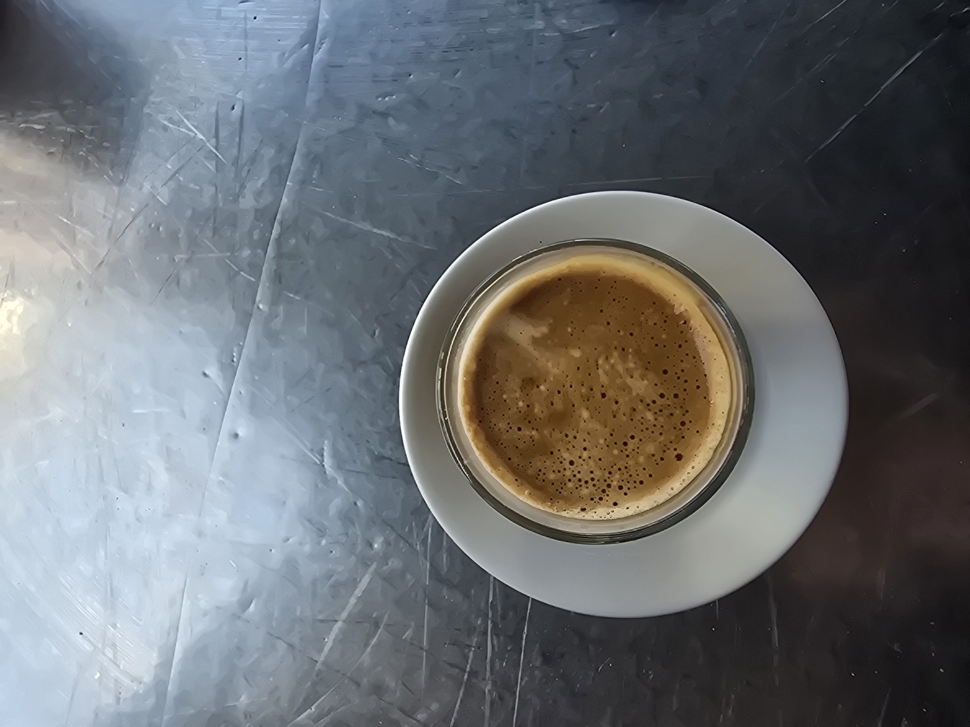 A top-down shot depicts a cup of coffee sitting on a white saucer. The cup is a transparent glass, revealing the brown, foamy coffee inside. The saucer is round and simple in design. The coffee and saucer rest on a reflective, brushed metal surface. The image was taken in Cornellà de Llobregat, Spain.