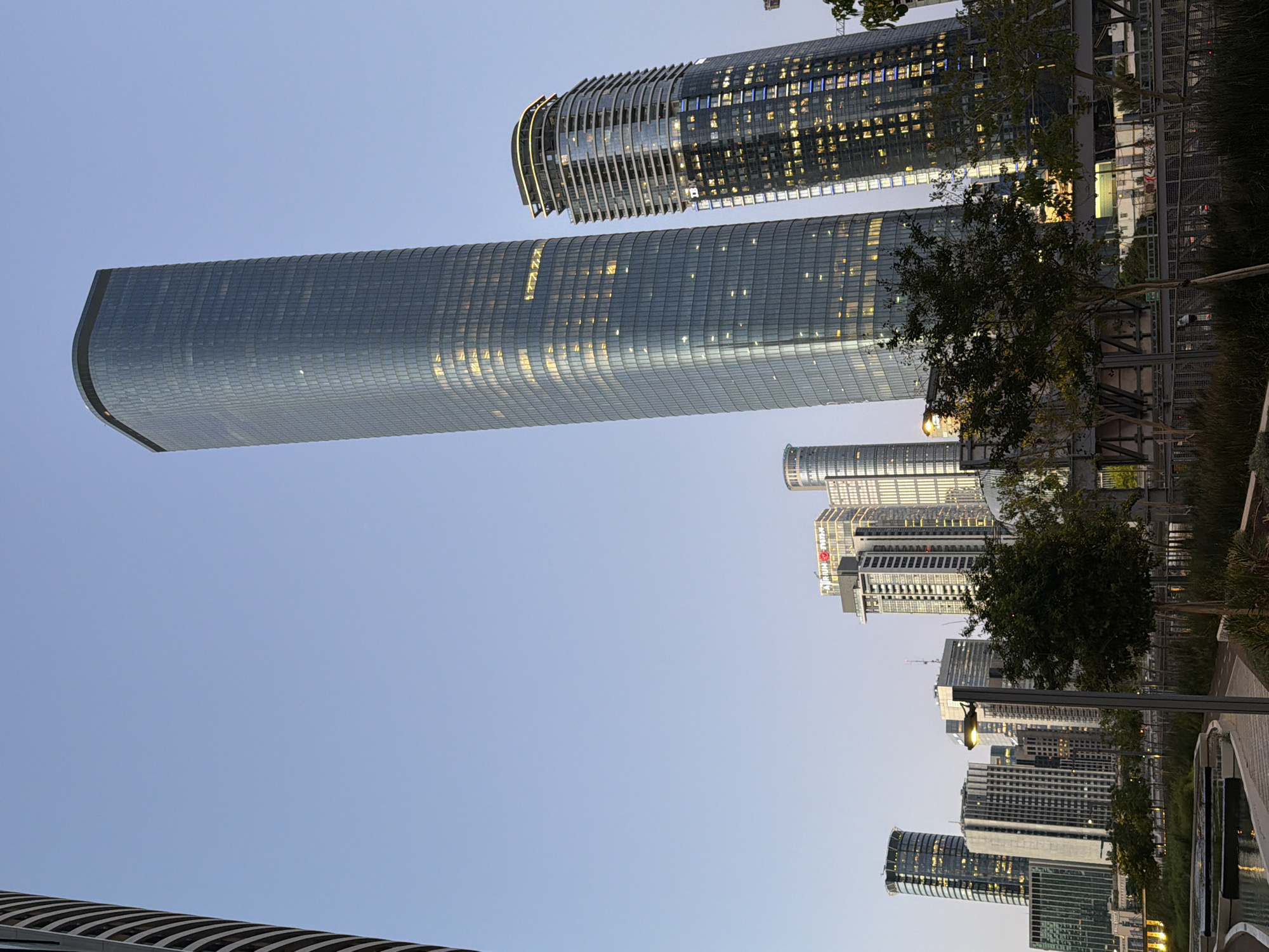 The image captures a skyline of modern buildings in Tel Aviv, Israel. Prominently featured are the Azrieli Sarona Tower, a long rectangular skyscraper, and the Azrieli Spiral Tower. Various other contemporary high-rise buildings surround these structures. Trees, and a paved walkway with built-in planters are also visible. The sky is a light blue, indicating either early morning or late evening. The image is rotated 90 degrees counterclockwise.