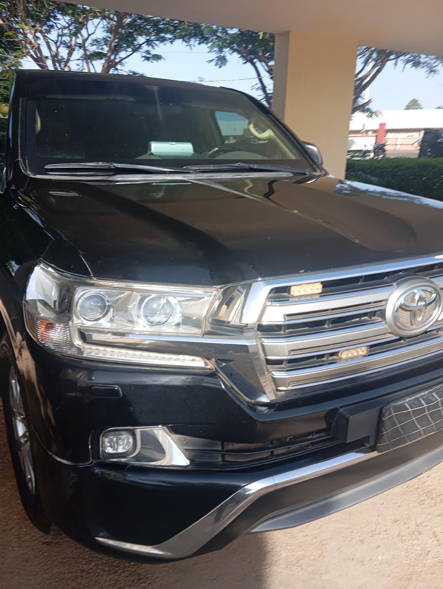 This media file shows the front of a black Toyota Land Cruiser parked outdoors. The vehicle is clean and appears to be in good condition. The front grille is prominent, featuring horizontal chrome bars and the Toyota emblem. Two rectangular amber lights are mounted on the grille, one on each side. The headlights are large and clear, with visible bulbs and reflectors. Below the grille, the bumper has a chrome accent and a black skid plate or cover on the right side. The vehicle is parked on a sandy or dirt surface, suggesting an outdoor environment. In the background, there are glimpses of trees with sparse foliage against a clear blue sky, and a portion of a building with a beige-colored pillar and a white roof is visible on the right side. Sunlight is casting shadows, indicating it is daytime. The overall scene is static, with the car being the primary focus. There is no visible text in the image. The context provided suggests the location is Jos, Nigeria.