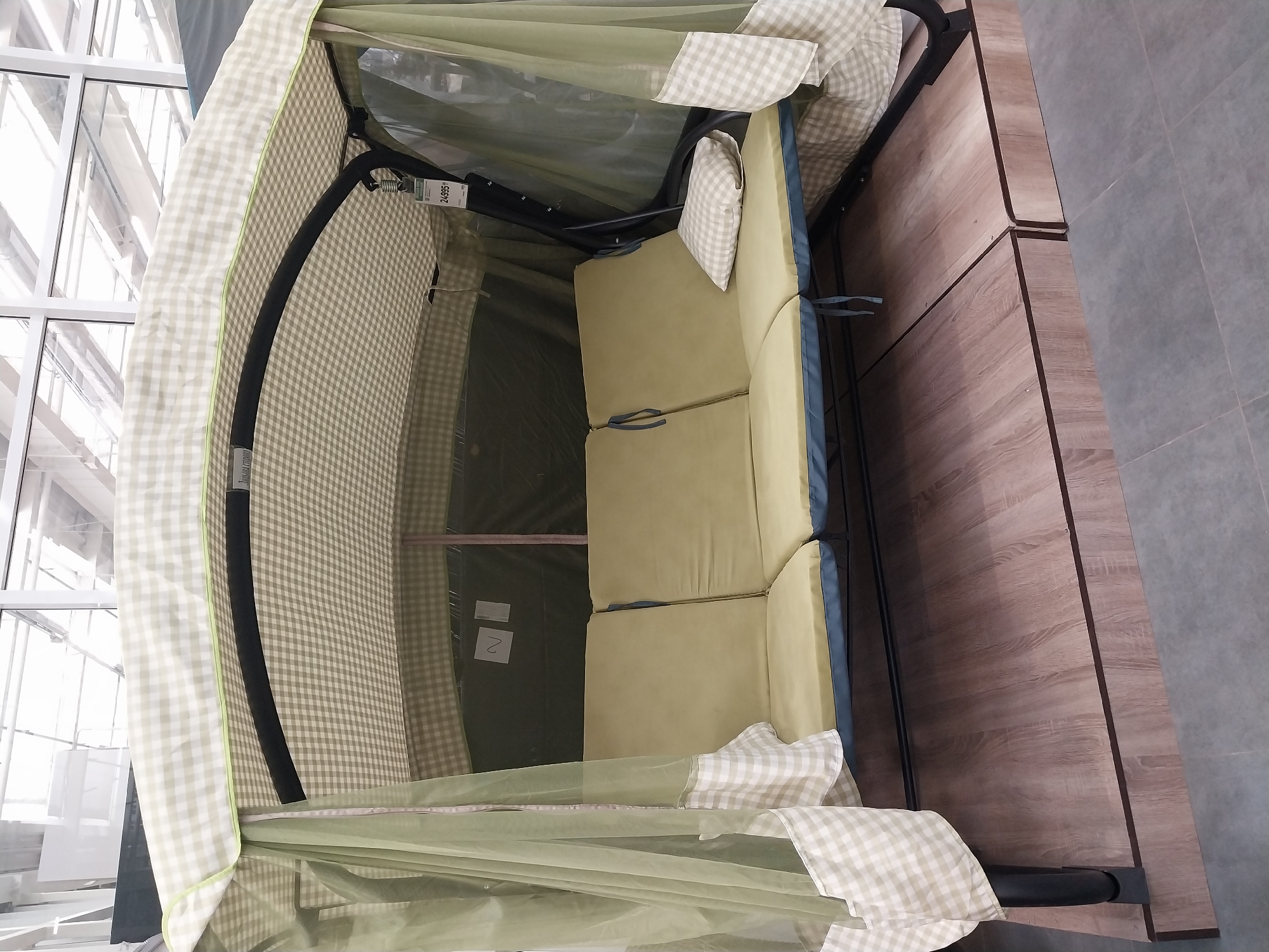 The image shows a garden swing set with a green and white checkered canopy and side curtains. The swing has a three-seat bench with light green cushions. A small checkered pillow rests on the right side of the bench. The swing is on a wooden platform, and behind it is a glass-windowed building with a visible ceiling structure. A price tag reading "24995" is attached to the swing's frame. The scene appears to be indoors, likely in a retail setting. The location is identified as Dnipro, Ukraine.
