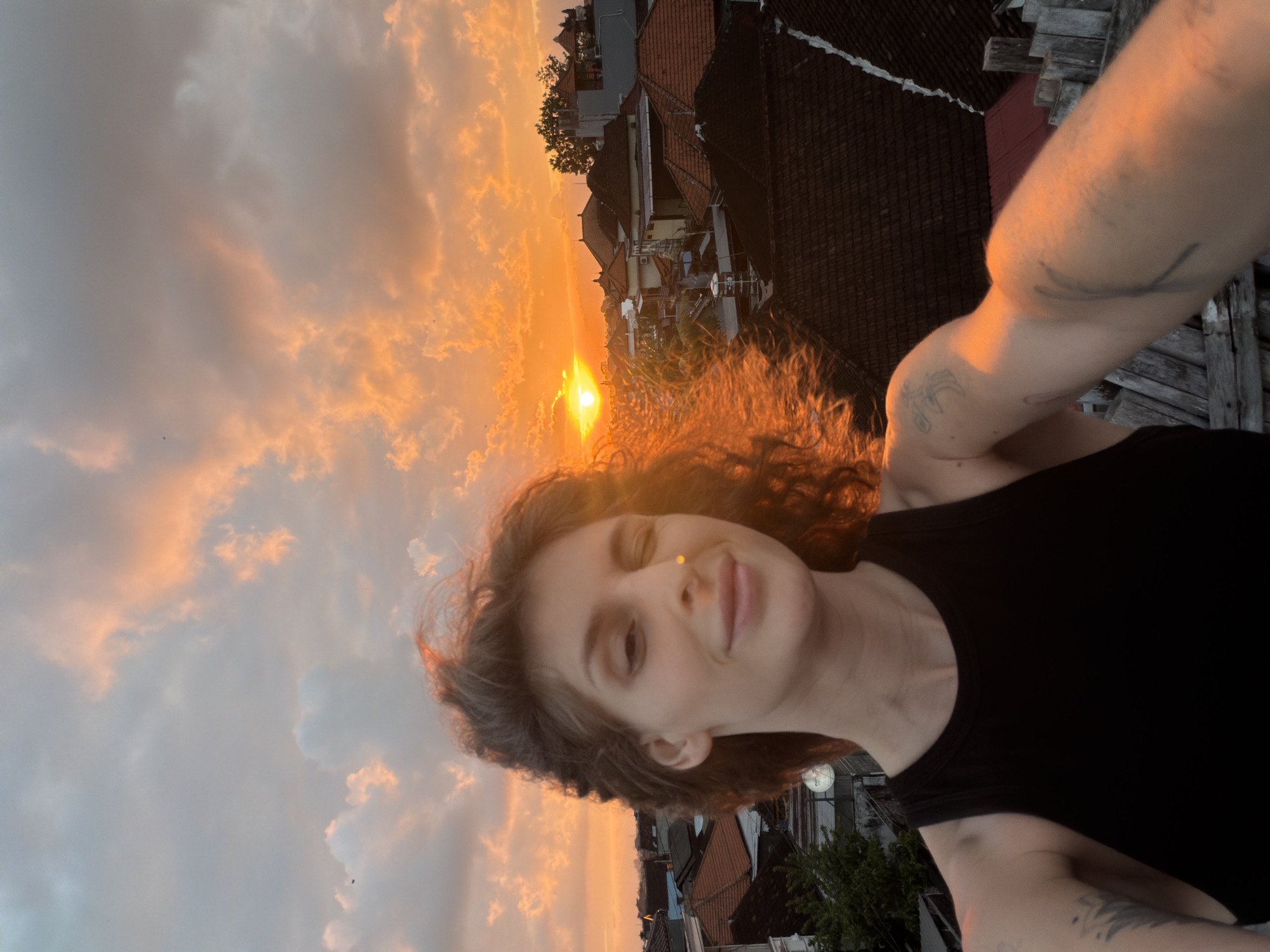 A selfie is taken from a rooftop in North Kuta, Indonesia, at sunset. The woman in the foreground is looking at the camera and winking. Her hair is windswept, and the golden light of the setting sun illuminates her face and arms. In the background, the roofs of houses and buildings are visible, with trees and the vibrant orange sky filled with clouds. The sun is setting on the horizon, casting a warm glow over the scene.