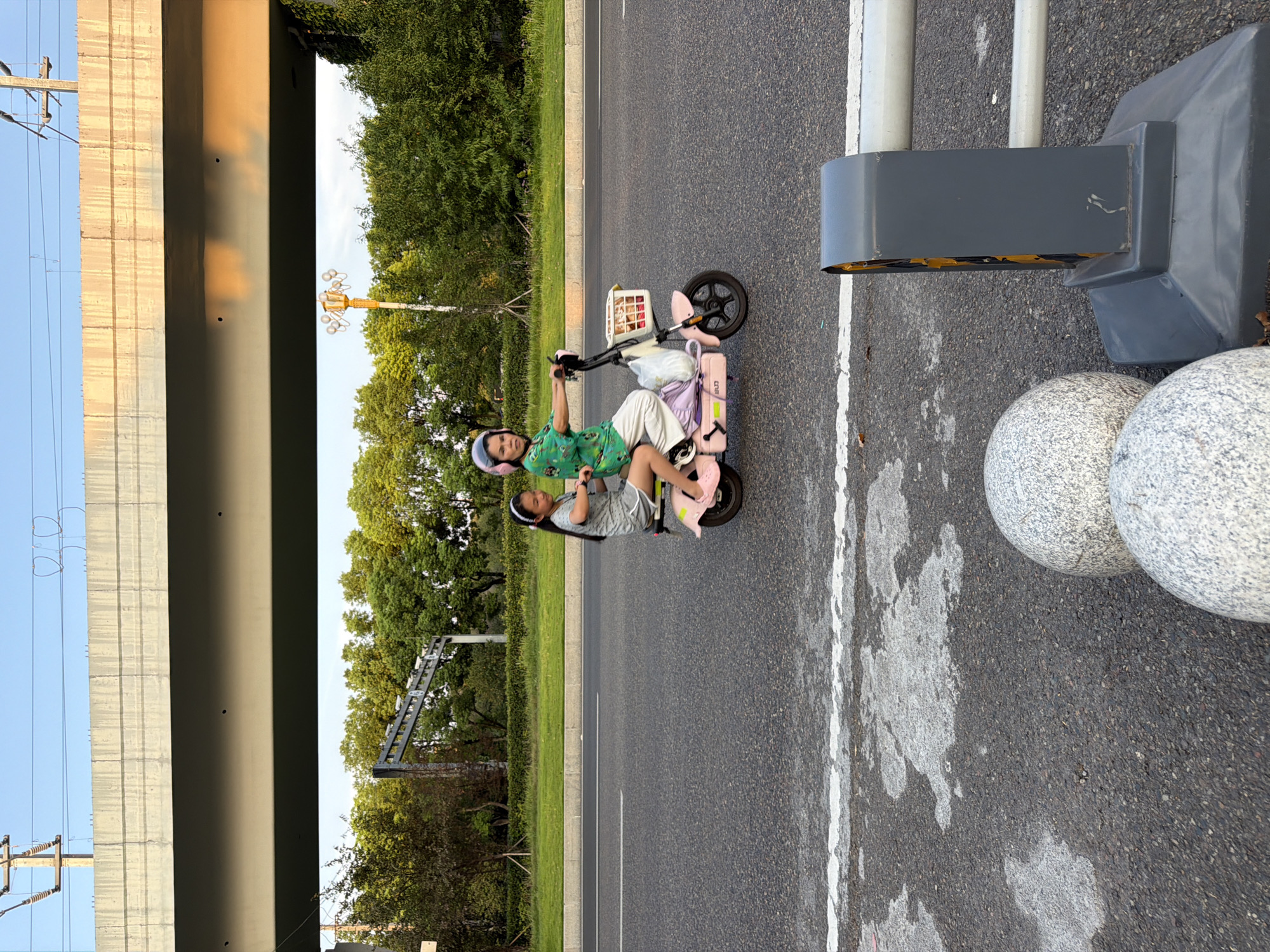 An adult woman wearing a green shirt, white pants, and a pink helmet rides a pink electric scooter down a paved road. A young girl, wearing a grey t-shirt and shorts, sits behind the woman on the scooter. The scooter has a white basket in front containing packaged food. A purple bag is attached to the rear of the scooter. The road is lined with a grassy median and trees. In the background, a concrete overpass stretches across the scene. The image appears to have been taken in the city of Jiangjin, China.
