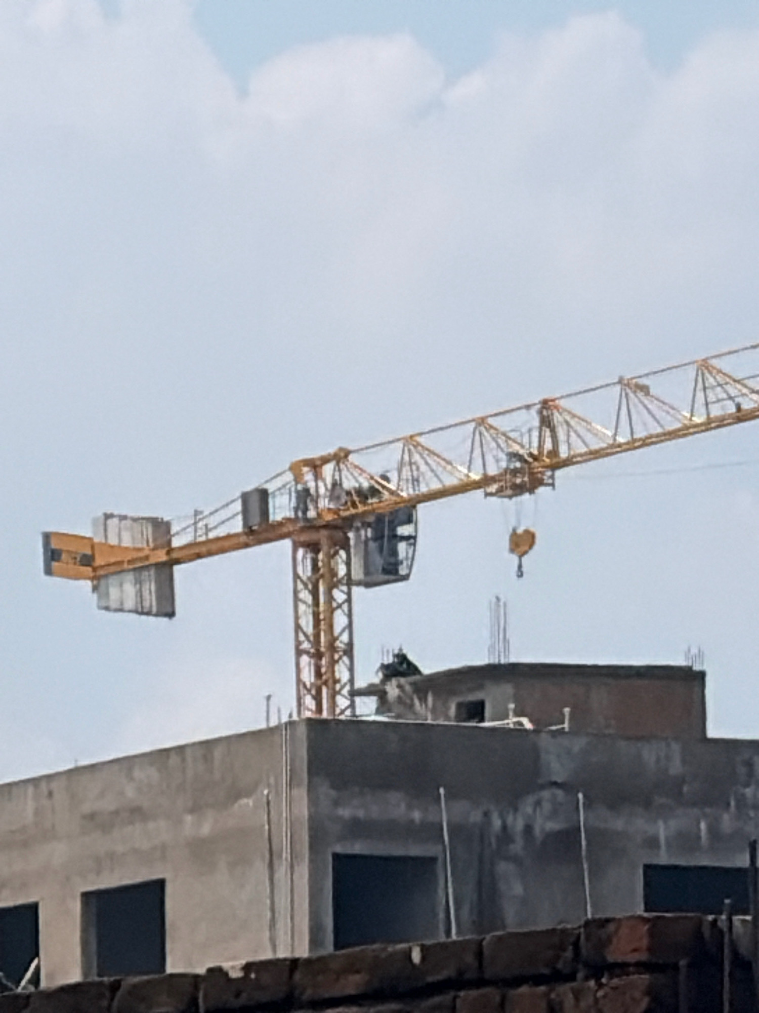 In Jamshedpur, India, a construction scene unfolds against a hazy, pale blue sky with scattered white clouds. Dominating the frame is a tall, yellow tower crane, its boom extending horizontally across the image. The crane's operational cab is visible, and a hook hangs from the boom. Below the crane, the partially constructed upper floors of a modern building are visible. This structure, made of concrete and featuring dark, empty window frames, suggests ongoing development. In the foreground, a brick wall and more unfinished construction elements contribute to the overall impression of a city in progress.