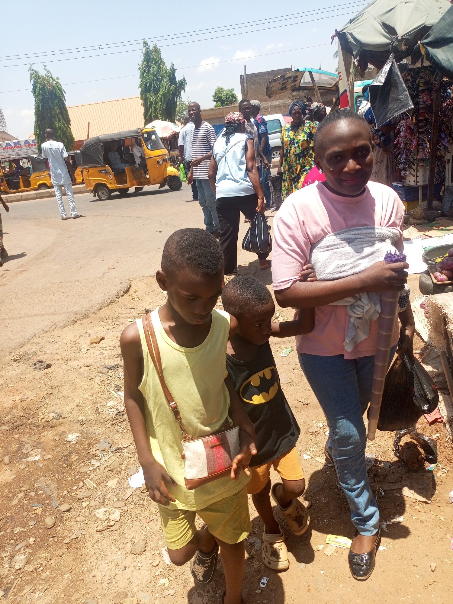 The image depicts a daytime scene in Sabon Tasha, Nigeria. In the foreground, a woman is walking with two young children. The woman is wearing a pink shirt and blue jeans, and she is carrying a baby wrapped in a white cloth. One child wears a yellow tank top and shorts, with a small purse strapped across their body, while the other wears a black Batman t-shirt and orange shorts.
The background shows a busy street with other people, including some wearing colorful traditional clothing. A yellow auto rickshaw, or "keke," is visible on the street, along with other vehicles. There's a stall selling items under a canopy, indicating a market or commercial area. The ground is a dusty, unpaved surface.