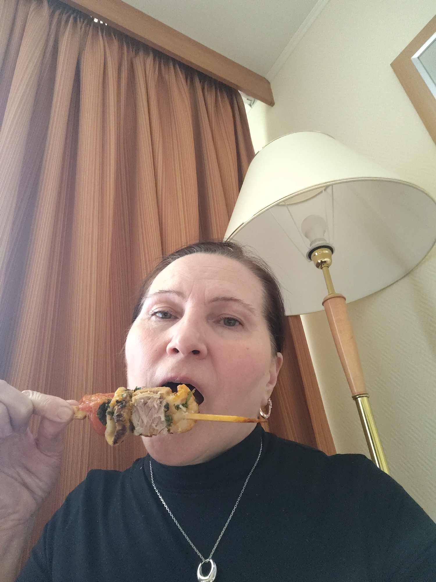 A medium close-up shot shows a woman indoors, appearing to take a selfie while eating from a skewer. She is positioned slightly off-center to the left, looking directly at the camera with her mouth open around a piece of food.
The woman has dark hair and is wearing a black high-neck top, a silver chain necklace with a circular pendant, and silver hoop earrings. She holds a wooden skewer with her left hand, which has pieces of grilled meat, possibly tomatoes, and green vegetables or herbs.
In the background to the left are pleated, vertically striped brown or tan curtains, hanging from a wooden rod near the top of the frame. To the right, a floor lamp with a cream-colored fabric lampshade and a visible light bulb dominates the upper right quadrant. The lamp's stem features a light-colored wooden section and a brass-colored base and fittings. The wall behind the lamp is a plain light cream or beige color. Part of a wooden frame, possibly a mirror or picture frame, is visible in the upper right corner.
The setting appears to be a room, possibly a hotel room, in Moscow, Russia. The light in the room is ambient, without strong natural light sources, and the lamp is either illuminated or the bulb is exposed.