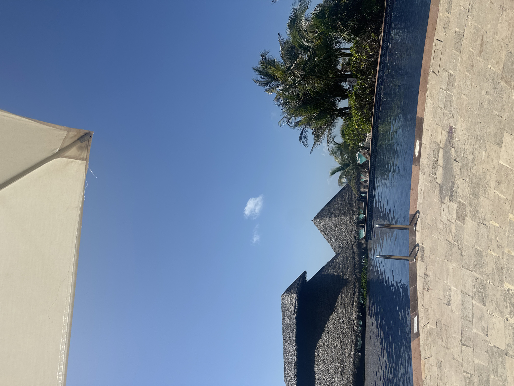 The image shows a daytime scene, likely taken in Cancún, Mexico, given the palm trees and architecture often associated with tropical resorts in that area. A white parasol is visible in the lower left corner. The sky is clear blue with a small white cloud. Two thatched-roof structures, possibly palapas, are positioned centrally. Adjacent to them, a dark-colored, still pool is lined with what appears to be a stone pathway and stairs leading into the water. Palm trees are visible along the side of the pool. The image is taken from a slightly low angle, and the camera is tilted. No people are visible in the image.