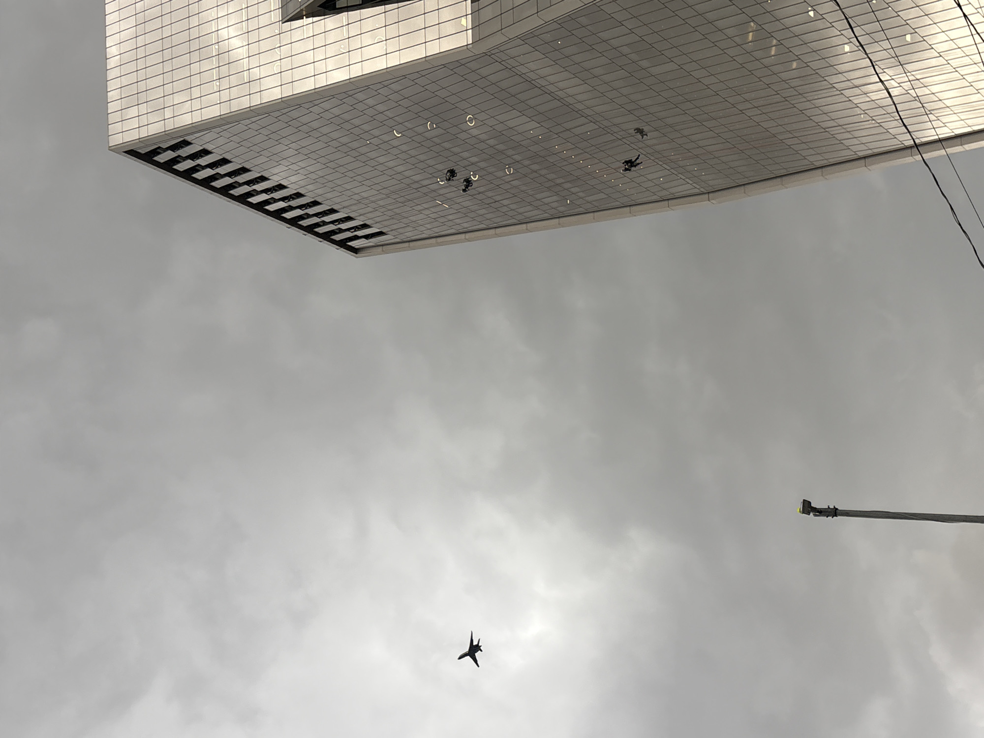 An airplane is flying in a cloudy sky above a modern glass skyscraper. Several window cleaners are rappelling down the side of the building. In the foreground, a street lamp is visible. The scene appears to be in a city. Based on the angle and architecture of the building, it is likely the Edifício Copan, designed by Oscar Niemeyer, located in São Paulo, Brazil.