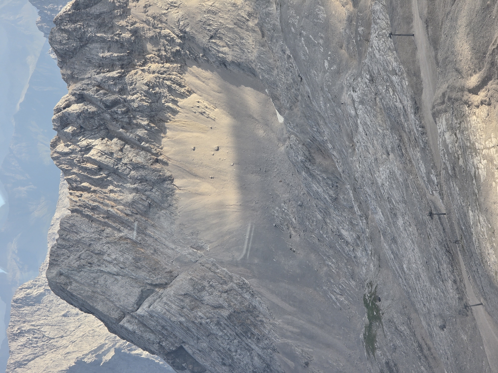 The image depicts a vast, rocky mountain landscape, likely the Zugspitze, the highest peak in Germany, located near Garmisch-Partenkirchen. The terrain is characterized by steep, rugged slopes covered in scree and exposed rock formations. In the foreground and midground, several ski lift pylons are visible, indicating a ski resort or an area accessible by cable car. The slopes are largely barren, with minimal vegetation, except for a small patch of green grass on a mid-level slope. The lighting suggests it is either early morning or late afternoon, with long shadows cast across the mountainside. The background shows a hazy expanse of further mountain ranges, fading into the distance. There are no visible people in the image. The overall scene conveys a sense of rugged natural beauty and a popular tourist or recreational destination.