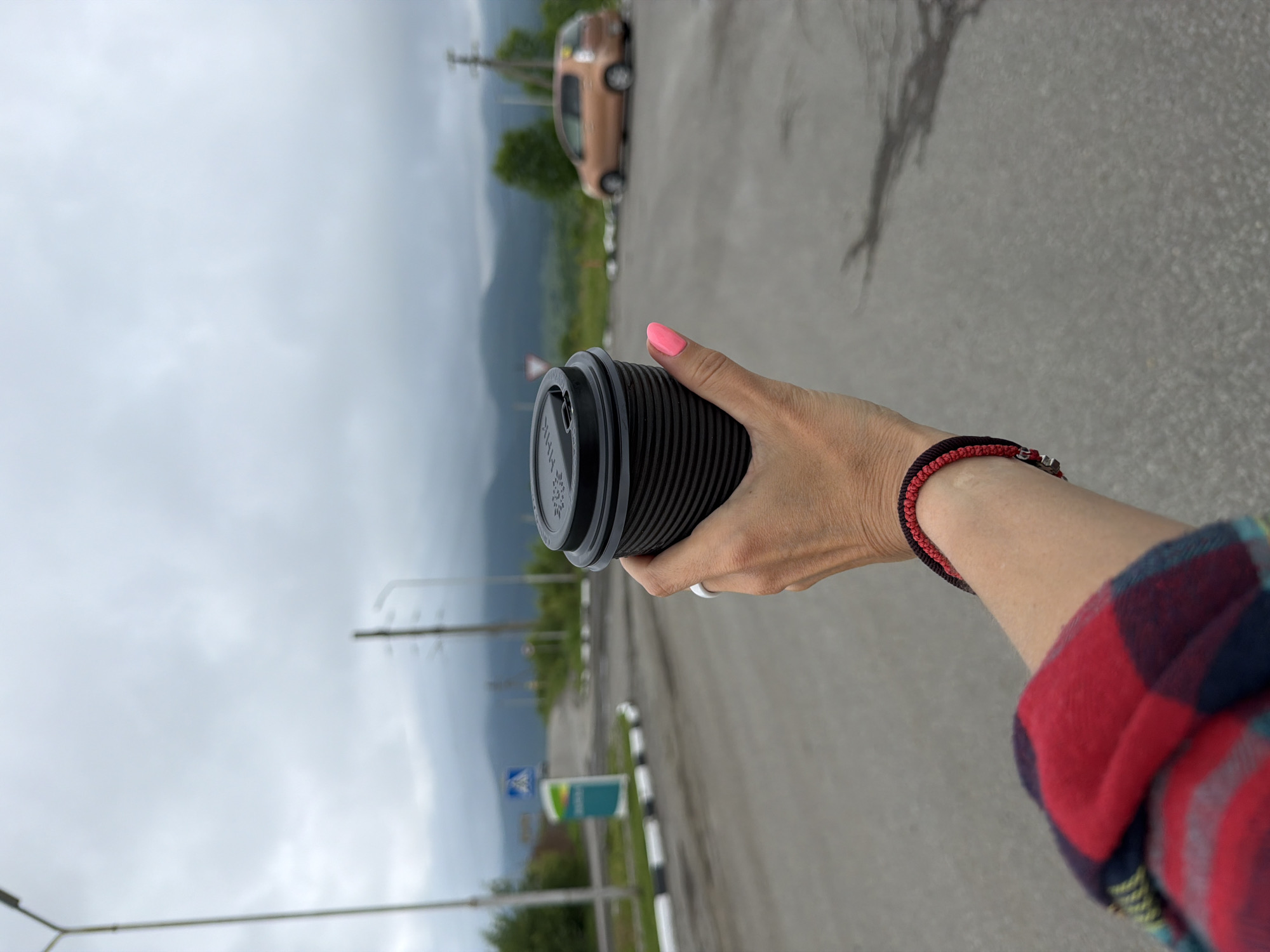 A person holds a black coffee cup in their right hand, with bright pink fingernails visible. They are wearing a red and blue plaid shirt and a braided red and black bracelet on their wrist. The background shows an asphalt road with a white and black striped curb. In the distance, there is a light brown car parked on the side of the road, and trees and mountains under a cloudy sky. Several street lights are visible in the background. A blue and green sign is present near the road. The scene appears to be outdoors in a rural or semi-rural setting. The time of day is likely daytime, given the visibility. The weather is overcast. The location context is Теплоозёрск, Russia. No discernible emotions are conveyed by the visible elements. There is text on the coffee cup lid and on the sign, but it is not legible.
