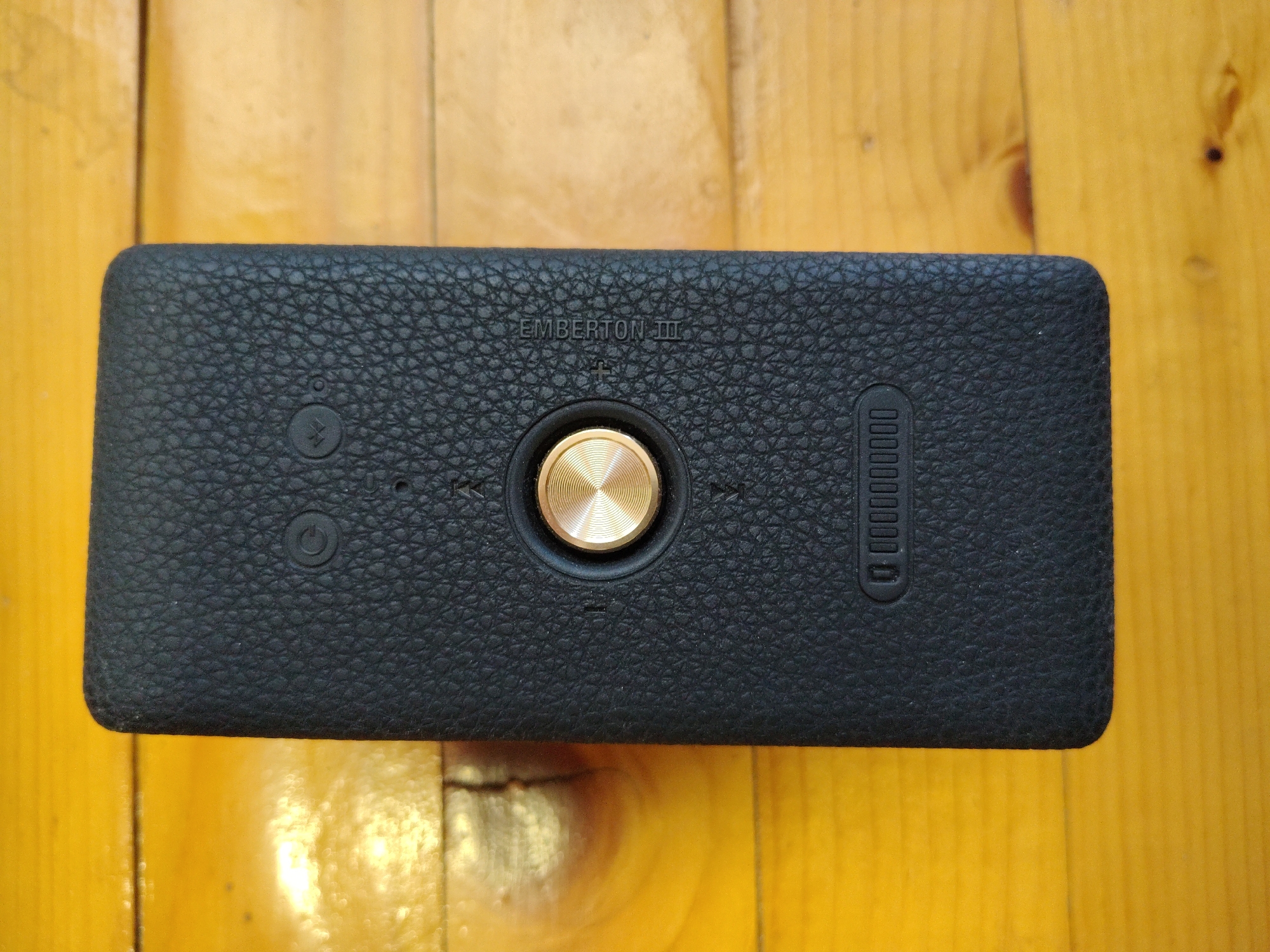 A black Marshall Emberton III speaker is placed horizontally on a light brown wooden surface. The speaker has a textured, faux-leather finish. On the top surface of the speaker, there is a prominent brass-colored control knob in the center, surrounded by smaller control buttons. To the left of the knob are two circular buttons, one with a power symbol and another with a lightning bolt symbol. To the right of the knob, there are play/pause and volume adjustment buttons. To the far right of the speaker, a series of vertical dashes and circles are visible, likely indicating battery status or connectivity. The wooden surface appears to be a table or countertop, with visible grain patterns and a slightly glossy finish. The lighting suggests an indoor environment, possibly with a light source casting subtle reflections on the wooden surface. The location is Unknown, Unknown.