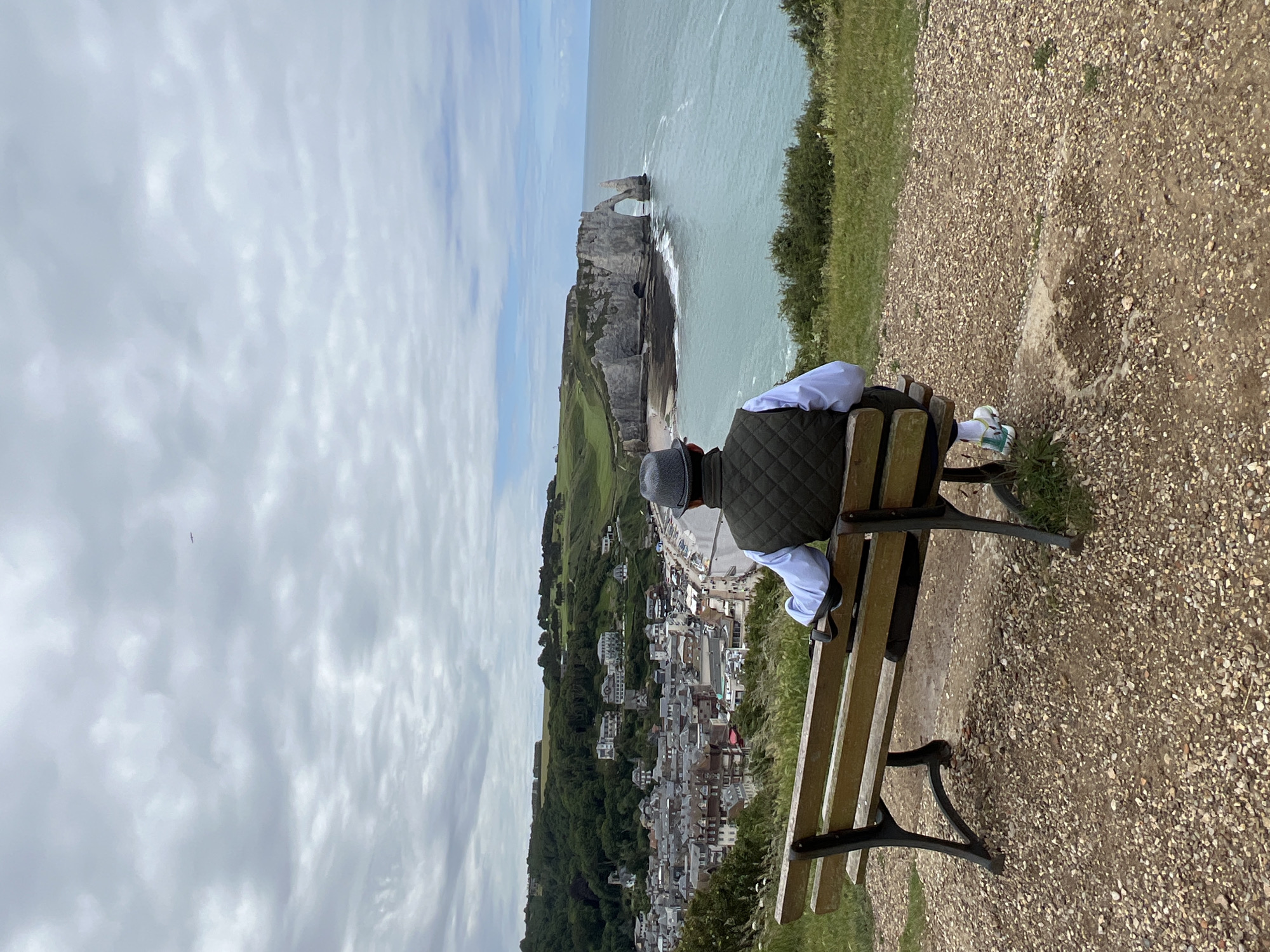 A person sits on a wooden bench, facing away from the camera, looking out at a coastal view. The person is wearing a dark quilted vest over a light-colored shirt and a grey hat. Their legs are crossed at the ankles, and they are wearing white and teal sneakers. The bench is made of wood with black metal legs and is situated on a gravel path overlooking a steep cliff.
In the background, a dramatic rock formation, known as the Falaise d'Aval, juts out into the turquoise ocean. The arch in the rock formation is clearly visible, and waves are breaking against the base of the cliffs. Below the cliffs and to the right, a charming town with many buildings is nestled in a valley, with green hills and trees surrounding it. The sky is partly cloudy, with patches of blue visible through the white clouds. The overall scene suggests a peaceful moment of contemplation and appreciation of natural beauty. The time of day appears to be daytime, possibly late morning or afternoon, given the lighting. The location is definitively Étretat, France, due to the iconic chalk cliffs and arch. There is no visible text in the image.