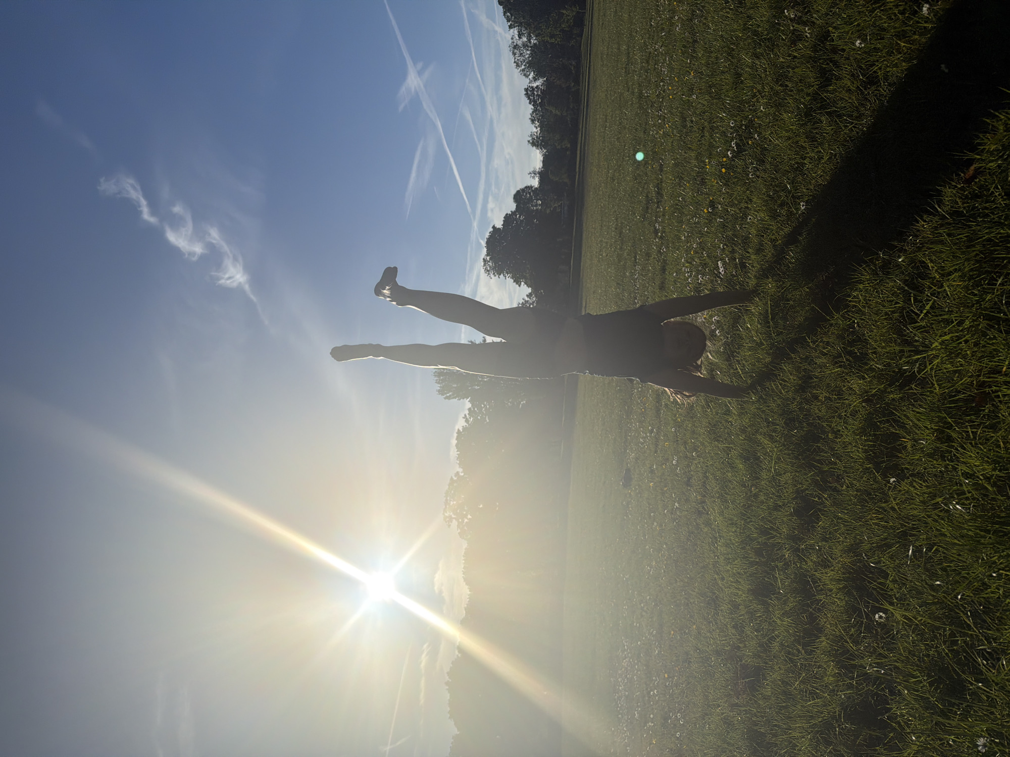 A person is positioned horizontally in a field, arms outstretched and legs extended upwards towards the left side of the frame. The person's body is silhouetted against a bright sun that is positioned at the bottom left corner of the image, creating a lens flare effect. Above the person, the sky is blue with contrails and some scattered clouds. The field is green with some small white flowers. A line of trees is visible in the background, adding depth to the scene. The image is oriented with what would typically be the right side of the picture facing upwards, giving the impression that the person is performing an acrobatic movement in the air. The location is likely a park with a large open grass area based on the surroundings. The city of London, United Kingdom cannot be identified from the provided image.