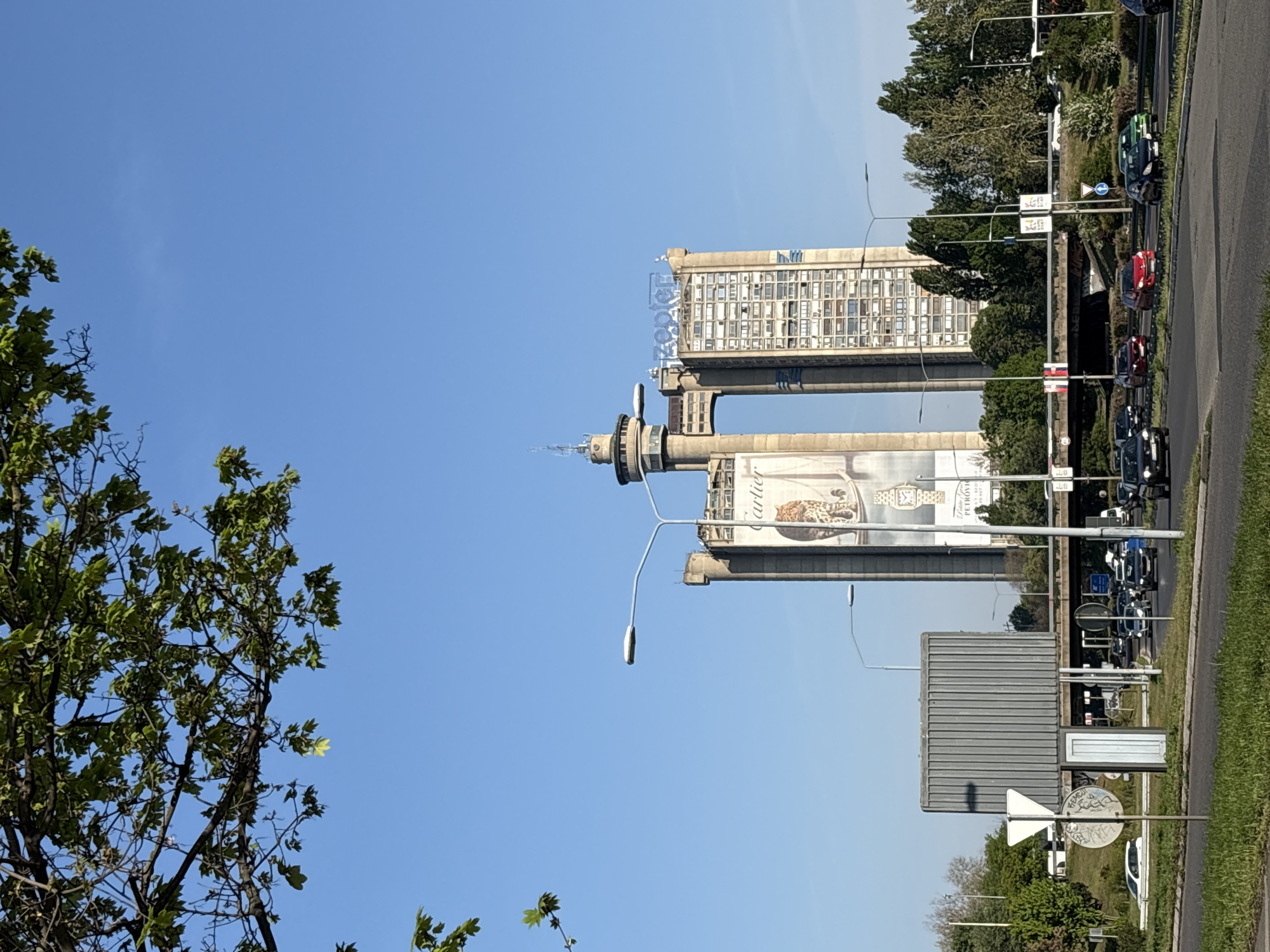 The image shows the West Gate (Genex) Towers of Belgrade, Serbia, viewed from street level on a clear day. The towers, comprising a residential tower and a commercial one, are connected by a two-story bridge and topped with a revolving restaurant. The residential tower displays "Genex" on its upper floors. An advertisement is visible on the commercial tower. A multi-lane road runs alongside the towers, with cars moving in both directions. Streetlights and signage are placed along the road. Green trees and vegetation are present on the roadside, and a tree is in the foreground on the left side. The sky is clear blue.