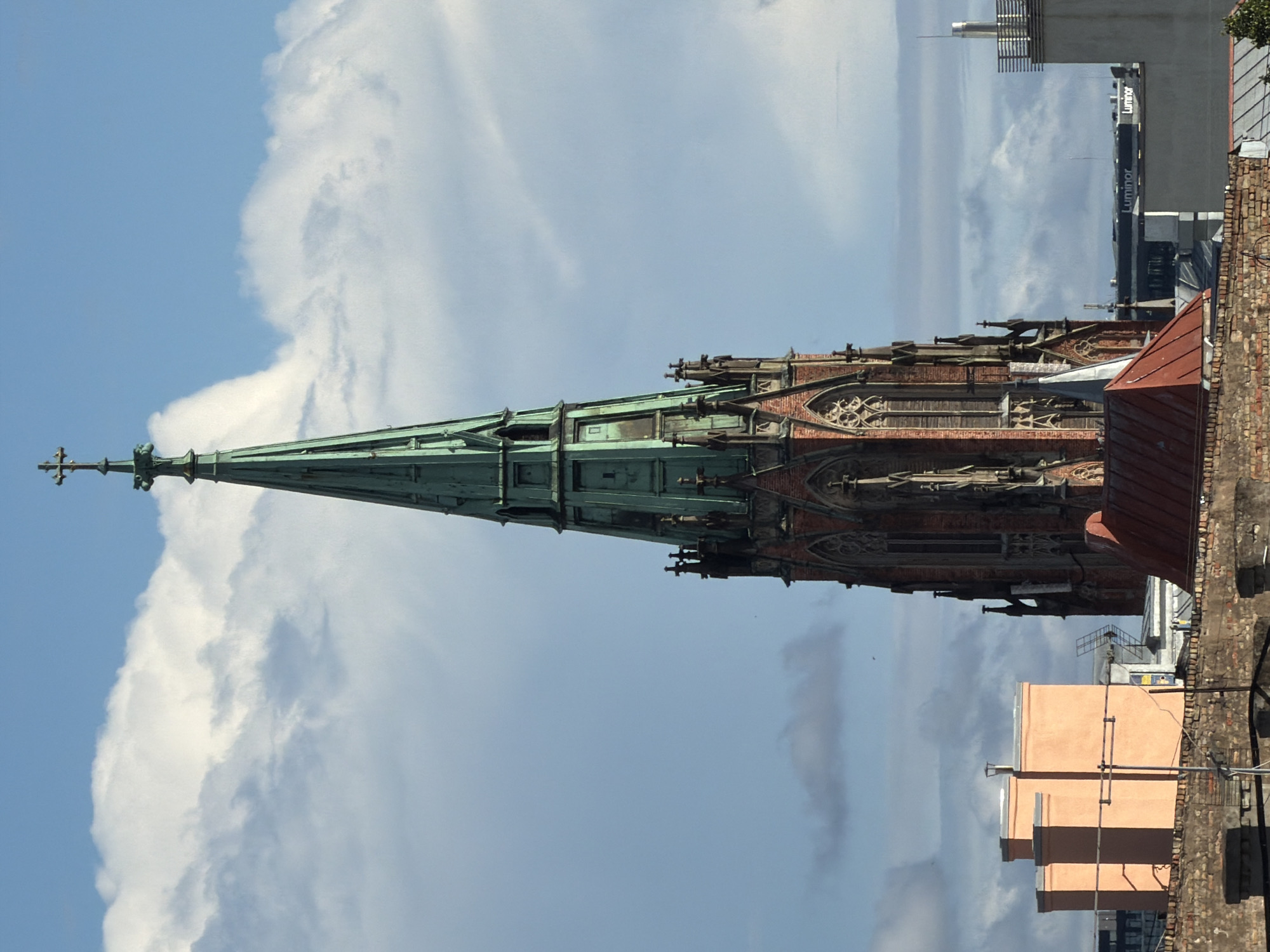 The image captures a section of the Riga city skyline on a partly cloudy day. The most prominent structure is a tall, Gothic-style church tower with a green, spire-shaped roof, topped by a cross. The tower is constructed of red brick and features intricate architectural details such as pointed arches and decorative gargoyles.
In the foreground, a brick wall forms the lower part of the scene, with parts of roofs visible beyond it. To the left of the church tower, a bright orange building with a flat roof and several chimneys stands out. Further in the background, other buildings and structures of varying heights and designs are visible, including a modern building with the "Luminor" logo.
The sky is a mix of blue sky and white, puffy clouds, with some lighter, wispy clouds further in the distance. No people are visible in the image. The overall impression is one of an urban landscape dominated by historic architecture.