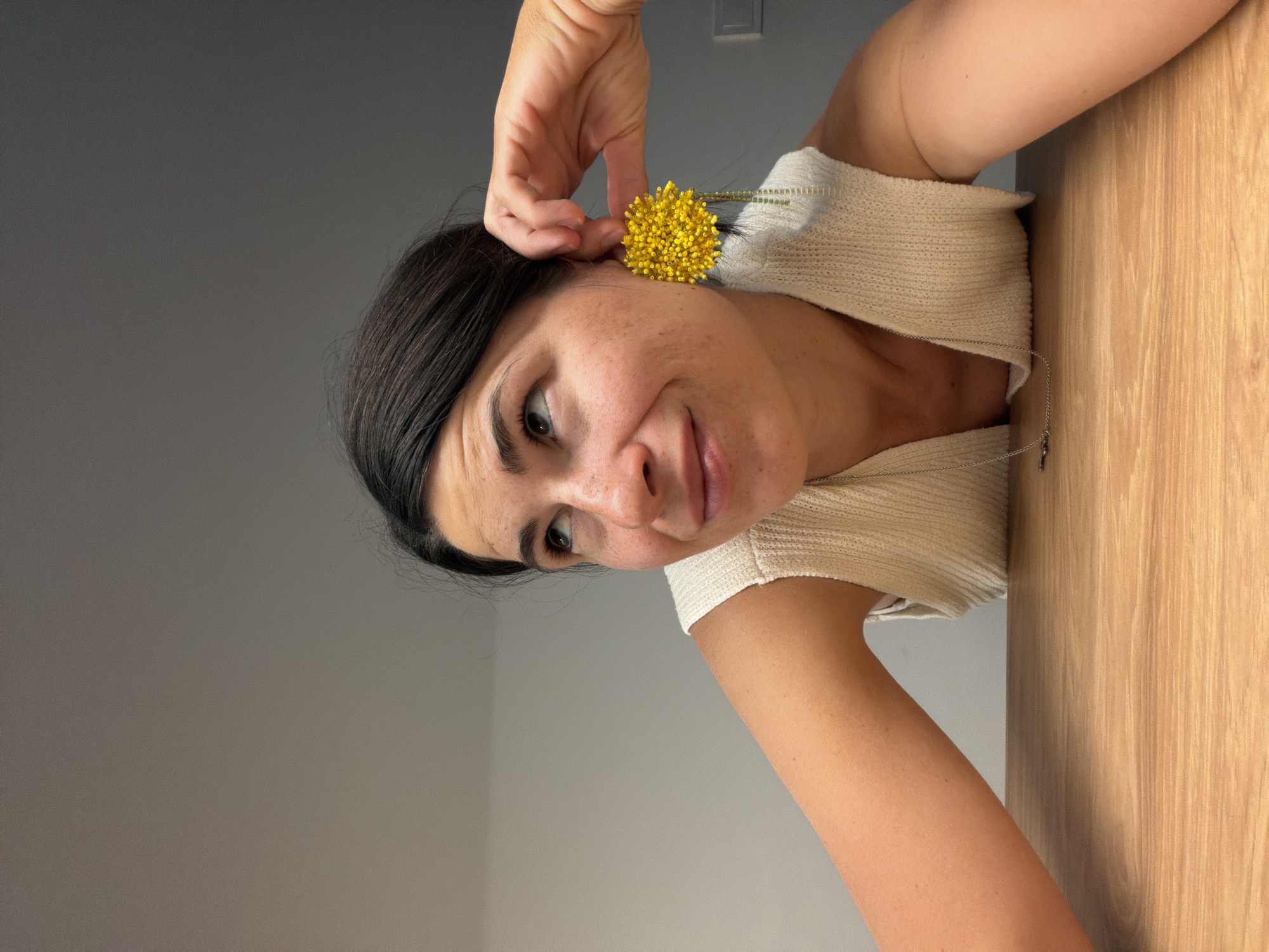 An indoor, portrait-style image shows a woman in an upright orientation, despite the original file's rotation. She is positioned towards the right side of the frame, looking to her left with a subtle smile and an amused expression. Her dark hair is styled around her face, which has fair skin and visible freckles. She wears a cream or off-white ribbed, short-sleeved top and a silver necklace with a small, dark-colored pendant.
Her right hand is raised, gently holding a bright yellow, spherical flower with a green stem near her left ear, as if placing it there as an adornment. The flower appears to be a variety of craspedia or similar bloom made of numerous small florets.
The background is divided into two distinct areas. To the woman's left and behind her is a plain, light gray wall, which takes up most of the left side of the composition. A rectangular light switch or outlet cover is visible on this gray wall in the upper portion of the frame. To her right, a light-colored wooden surface with a prominent vertical grain runs along the edge, suggesting a wooden panel, wall, or table. The overall lighting is soft and even, indicating an indoor setting. There is no visible text within the image. The context provided indicates the location is Budva, Montenegro.