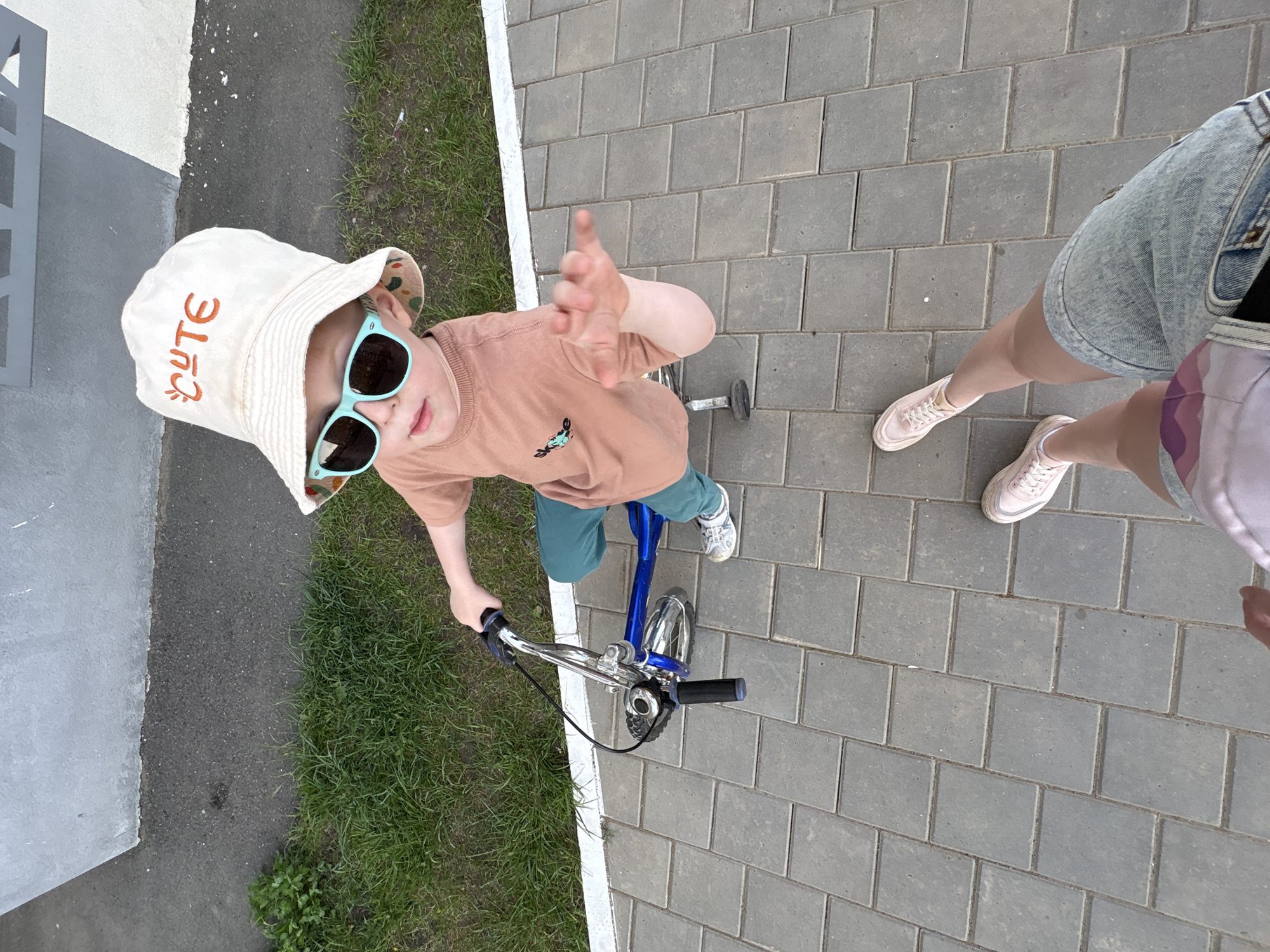 A high-angle photograph shows a child on a blue bicycle, likely a strider bike, with an adult nearby. The child wears a beige bucket hat with the word "CUTE" embroidered on the brim, a brown shirt with a small graphic, blue shorts, and sneakers. They are also wearing blue-framed sunglasses and have one hand raised, seemingly waving. The adult, partially visible on the right, wears denim shorts and pink sneakers. The scene takes place on a paved area composed of gray rectangular bricks. A patch of grass is visible to the left. The location is Izhevsk, Russia.