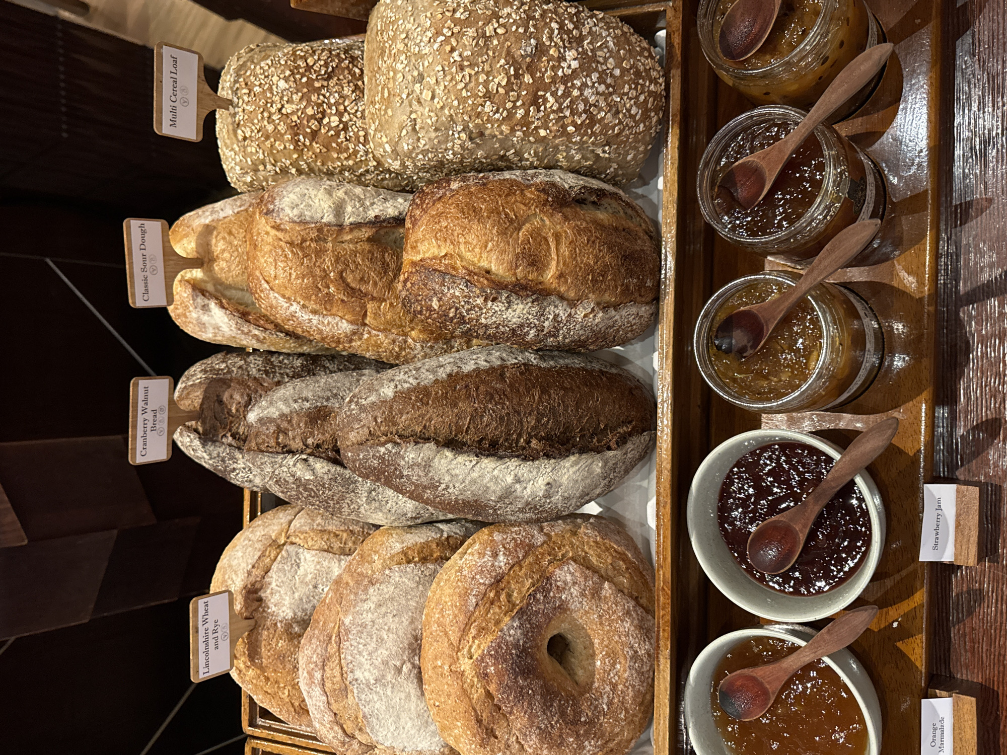 In Kuta Selatan, Indonesia, on a wooden tray, a selection of artisan breads and jars of jam are displayed. The breads, in various shapes and sizes, include loaves labeled "Lincolnshire Wheat and Rye," "Cranberry Walnut Bread," "Classic Sour Dough," and "Multi Cereal Loaf." Each bread appears rustic, with floured crusts and visible scoring. Below the breads, three small bowls and three glass jars contain different varieties of jam. The bowls hold jams that appear to be orange marmalade and strawberry jam, with wooden spoons resting in them. The glass jars also contain jams, with wooden spoons inserted into each. The overall scene is a well-presented breakfast or buffet spread.