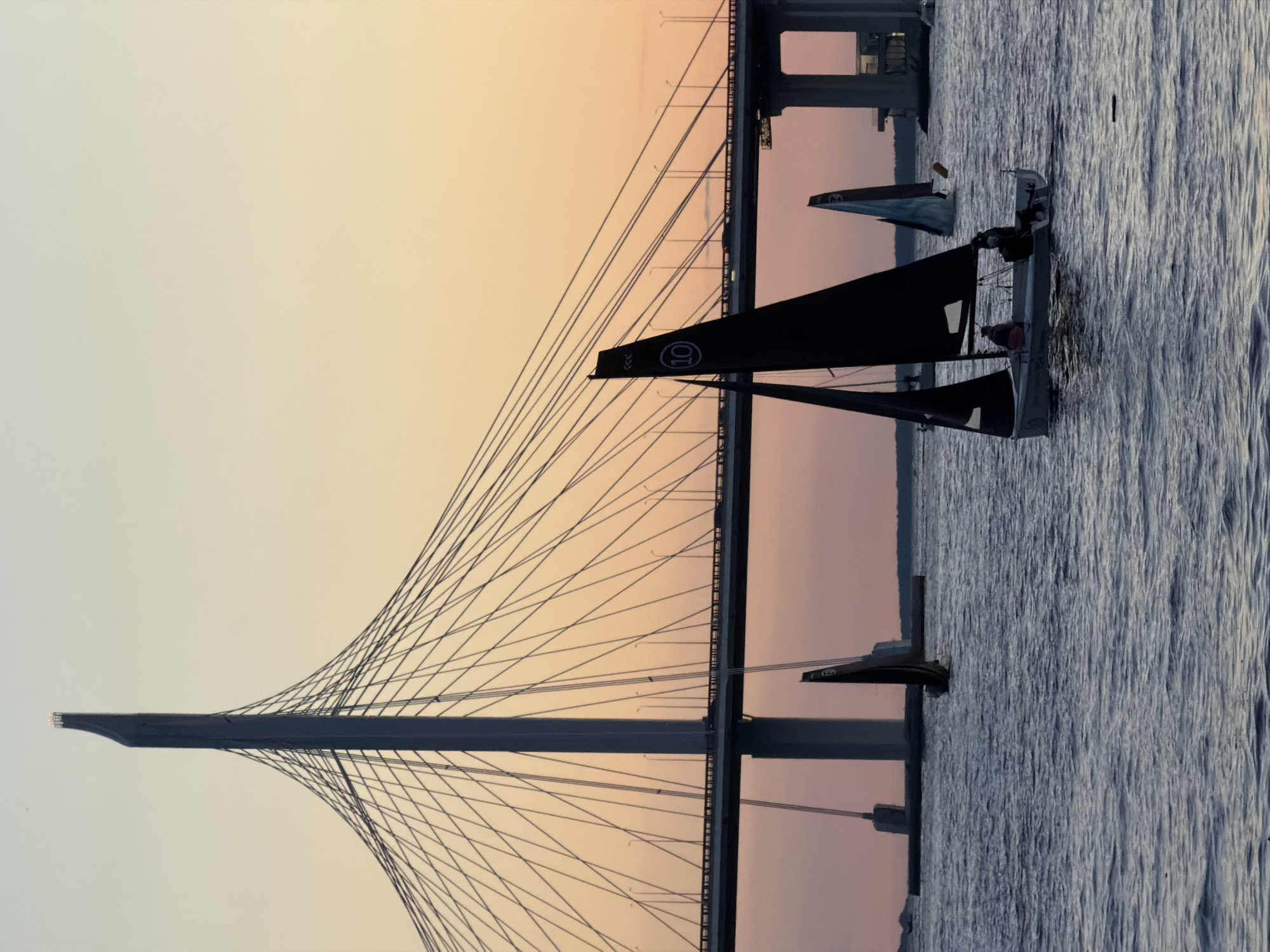 The image captures a scenic view of sailboats navigating on the water with a prominent cable-stayed bridge in the background. The sky exhibits a gradient of soft orange and pale yellow hues, indicative of a sunset or sunrise. The bridge, a modern architectural marvel, features a tall, slender pylon from which numerous cables extend to support the roadway. The water is choppy, reflecting the ambient light.
In the foreground, a dark-sailed sailboat is prominently featured, with two individuals visible onboard. Another sailboat with a lighter sail is positioned further back, and a third, smaller sailboat is also visible in the distance. The overall atmosphere is serene and tranquil, with the natural beauty of the seascape complemented by the man-made structure of the bridge. The scene is likely set in a city where such a bridge and waterway exist, possibly a port city or one situated on a large river or bay. The presence of sailboats suggests a recreational or sporting event taking place on the water.