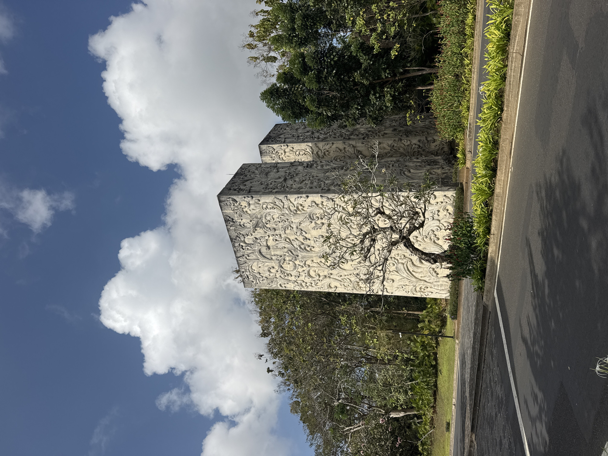 In Kuta Selatan, Indonesia, two imposing, rectangular stone structures stand side-by-side, adorned with intricate relief carvings of swirling floral patterns. These structures are partially obscured by lush green trees and foliage. In front of them, a bare-branched tree with a gnarled trunk stands out against the textured walls. A paved road runs in the foreground, with a white line marking the edge. The scene is set against a bright blue sky dotted with fluffy white clouds, suggesting a sunny day.