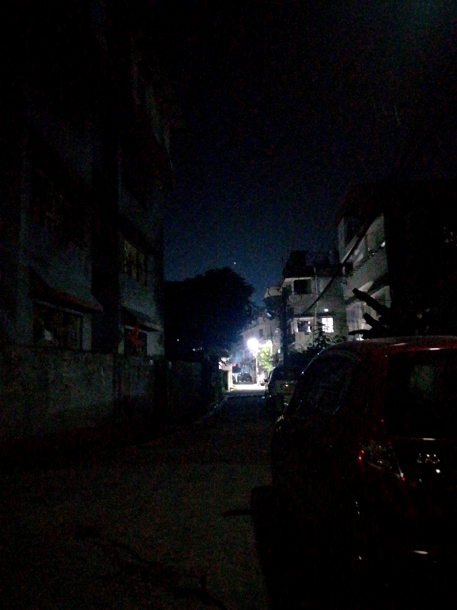 An image taken at night shows a street scene in Siliguri, India, with buildings and a parked car. On the left side of the frame, a multi-story building with a textured, dark facade is visible. Some windows on this building appear to have lights on. The ground in front of this building is paved and dark. In the center of the image, a narrow street recedes into the distance, flanked by more buildings and trees. Streetlights illuminate parts of the street and the facades of some of the buildings, casting a dim glow on the surroundings. A dark-colored car is parked on the right side of the street, partially obscuring the view of the buildings behind it. The sky above is dark, indicative of nighttime.
