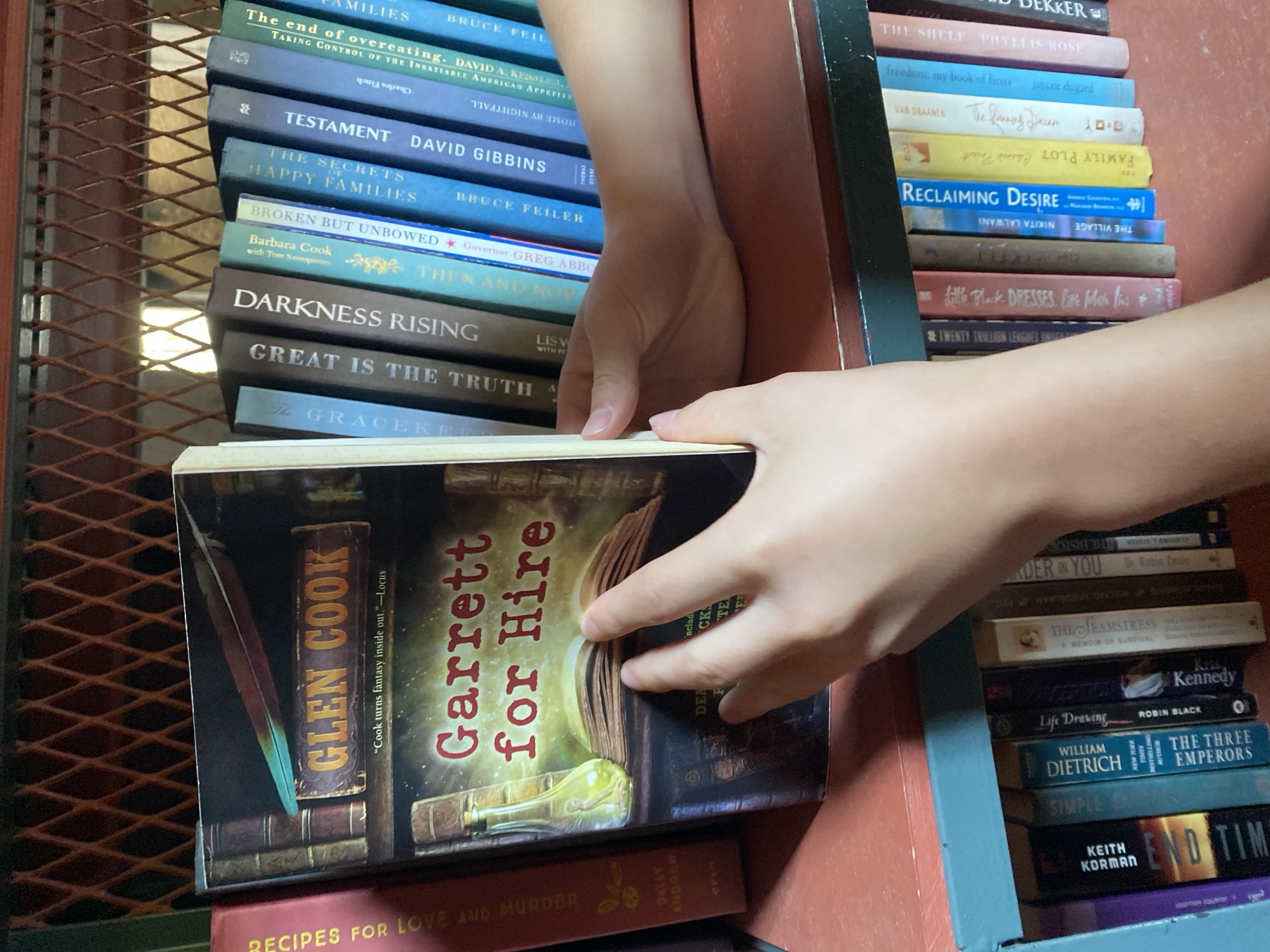 This image depicts a close-up view of a bookshelf filled with books, with a person's hands interacting with the books. The shelf is made of dark metal with a diamond-shaped pattern. The books are of various sizes and colors, with titles visible on their spines. A prominent book in the foreground has the title "Garrett for Hire" by Glen Cook. The person's hands are reaching for and touching the spine of this book, as well as another book to its right. The lighting suggests an indoor setting, possibly a library or a private collection. The lower shelf also contains a row of books with visible titles such as "END OF TIME" and "THE THREE EMPERORS." The overall impression is one of a casual interaction with books on a shelf. There is no information within the image to suggest a specific location in Tbilisi, Georgia.