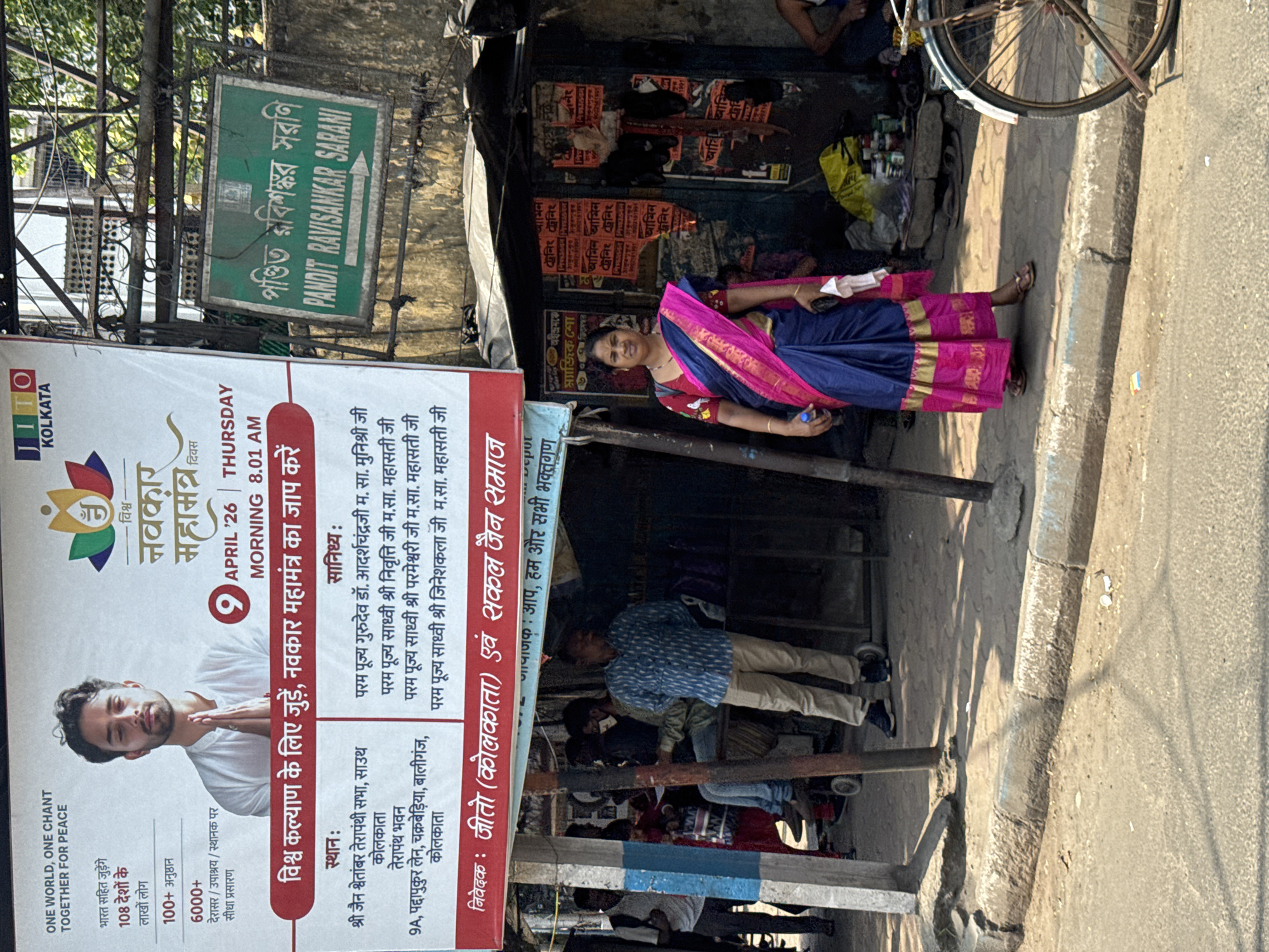 Here is a detailed description of the image:
**Overall Scene:**
The image shows a street scene in Kolkata, India. There is a banner advertising a "Navkar Mahamantra" event with the date of April 26th and a morning time of 8:01 AM. People are standing around, and a woman in a traditional sari is present. Overhead street signs are also visible.
**Banner Details:**
The banner features an image of a man and the event title "Vishva Navkar Mahamantra." It mentions "One World, One Chant Together for Peace" and lists statistics like "108 countries," "100+ programs," and "6000+ temples/sites." The banner also provides location details, including "Shri Jain Shwetambar Terapanthi Sabha, South Kolkata" and "Terapanth Bhavan, 9A, Paddapukur Lane, Chakraberia, Ballygunge, Kolkata."
**People:**
A woman in a blue and pink sari stands towards the middle right of the image. There are other people milling around near the banner and along the street.
**Objects:**
A sign that reads "Pandit Ravishankar Sarani," a Bengali street sign, posters on the wall behind the woman in a sari, and a bicycle wheel are visible near the top of the image.
**Location:**
The location is confirmed to be Kolkata, as indicated on the banner. The presence of Bengali text on the street sign reinforces this.
**Environment:**
The street appears to be a busy urban area with shops and pedestrians.