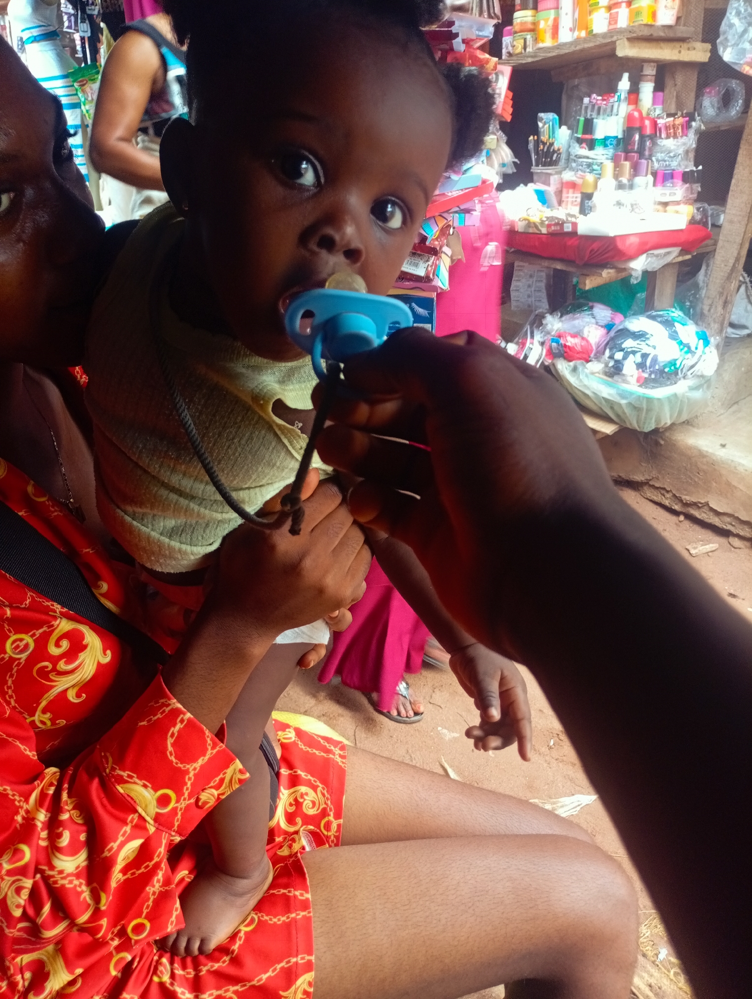 In this medium shot, a baby is being held and given a pacifier. The baby is of African descent and has dark hair and eyes. They are wearing a light green bib and are being held by a person whose arm is visible in the foreground. The person is wearing a red shirt with a gold chain pattern. In the background, shelves are stocked with various items, including cosmetics, toiletries, and other small goods, suggesting a market or shop setting. Sunlight is visible, indicating it is daytime. The overall scene appears to be outdoors or in an open-air market.