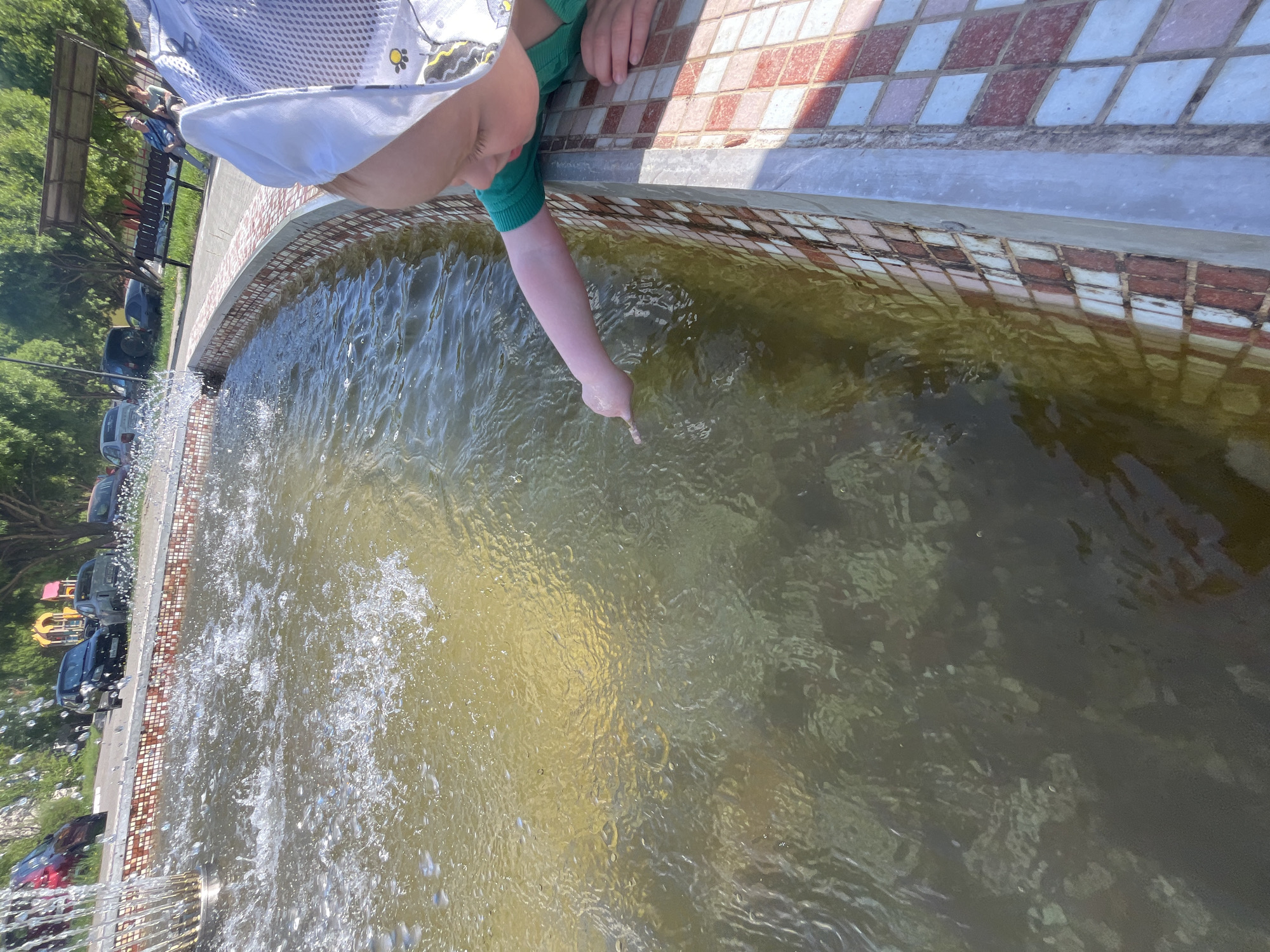 A child is interacting with a fountain, most likely in Chaikovsky, Russia. The child, dressed in green sleeves and a white shirt, is reaching into the murky water of the fountain with their right hand, index finger extended. The fountain is adorned with brick-patterned tiles along its edge. In the background, a park scene unfolds with trees, benches, cars, and play equipment visible along the street. The fountain's water jets are spraying upwards and outwards, adding motion to the scene.