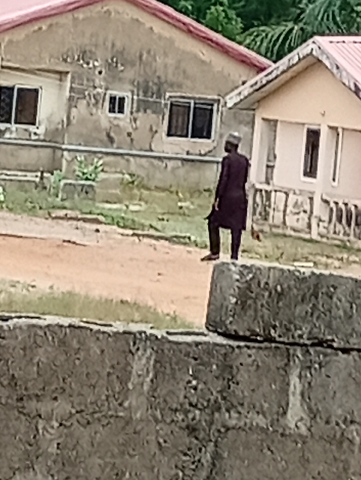 A man in a dark robe and hat walks across a dirt road in front of two buildings. The buildings have red roofs and windows. In the foreground is a concrete wall. The location is Unknown, Unknown.