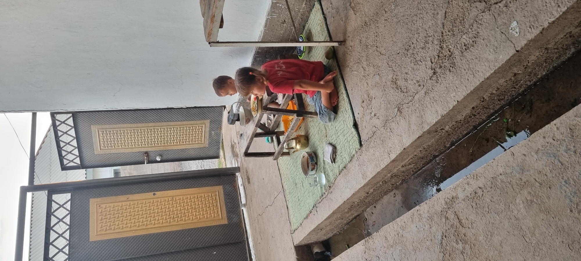 In Ҷомӣ, Tajikistan, two young children are seated on a green mat on a concrete ground near a drainage ditch. A young boy, facing left, is sitting with his legs tucked underneath him. A young girl, wearing a red t-shirt and denim shorts, is sitting on the mat facing right, with her legs crossed. In front of them, a low wooden table holds bowls of food and a metal teapot. The background shows a large double door with a textured design. A white wall is to the right of the door. The scene appears to be outdoors, likely in a residential area, with the concrete and drainage ditch suggesting a practical, functional outdoor space.