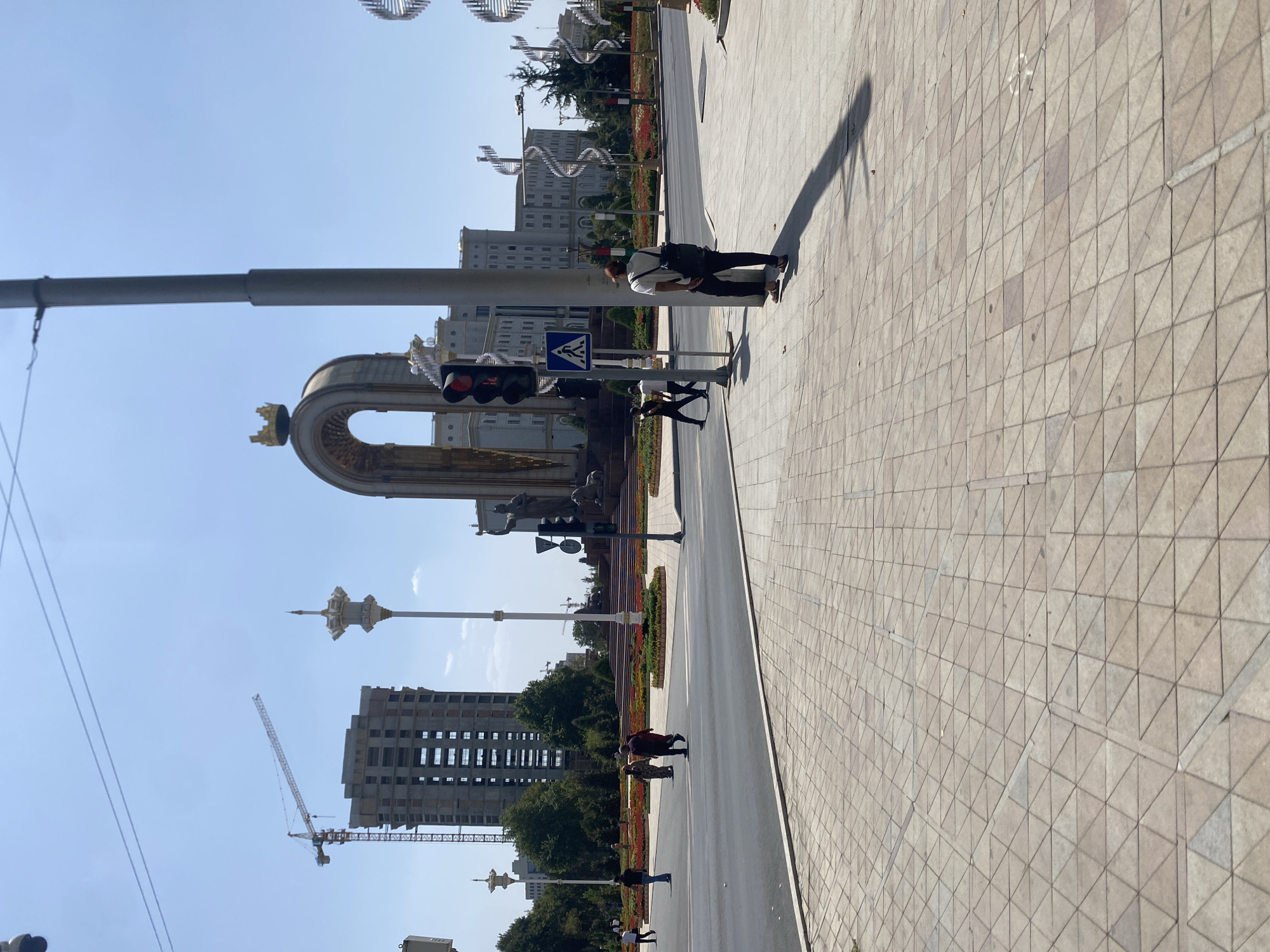 The image captures a bright, sunny day in Dushanbe, Tajikistan, likely at the prominent Rudaki Park. In the foreground, a man with a backpack stands on a paved sidewalk, casting a long shadow. Across a wide, multi-lane street, a large, ornate archway structure, topped with a golden crown, dominates the mid-ground. This landmark, likely the Arch of National Unity, is surrounded by lush greenery and flower beds. Adjacent to it, a grand, multi-story white building with arched windows stands prominently. Further in the background, a partially constructed tall building and a construction crane indicate ongoing development. Several individuals are visible crossing the street at a designated crosswalk, with traffic lights indicating the flow of movement. The environment suggests a bustling urban center with significant architectural and public spaces.