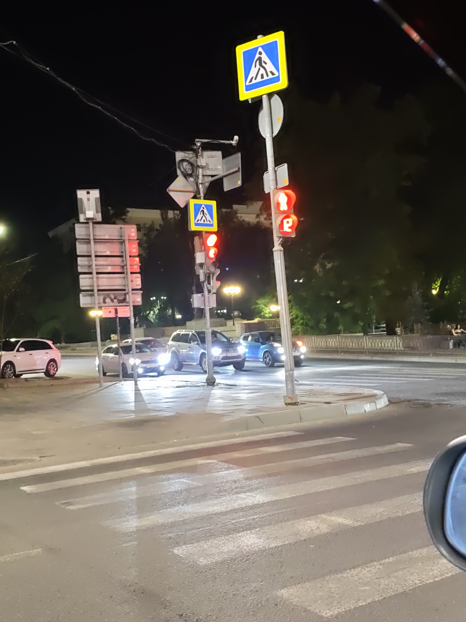 The image depicts a nighttime street scene at an intersection with a pedestrian crossing in Rostov-on-Don, Russia. Traffic lights are visible, with the red light illuminated for pedestrian signals, indicating that pedestrians should not cross. A pedestrian crossing sign, depicting a person walking, is mounted on a pole above the traffic lights. Several cars are present at the intersection, including a white SUV and a blue sedan. The street surface appears wet, suggesting recent rain or street cleaning. Trees and streetlights are visible in the background, contributing to the urban nighttime ambiance. There are no discernible people actively crossing the street at this moment. The context suggests a typical urban environment during evening hours.