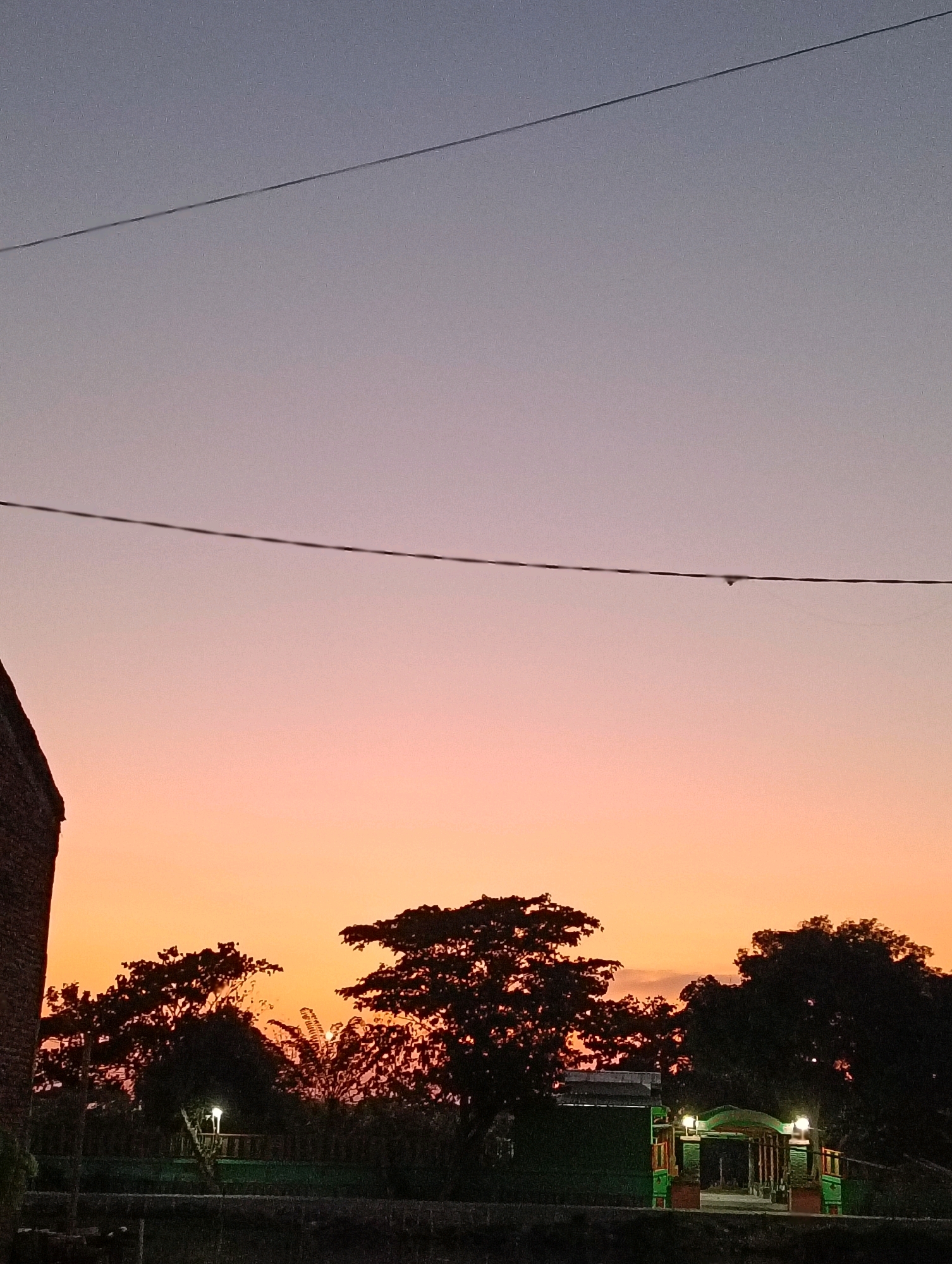 This is a photograph of a sunset in Semarang, Indonesia. The sky is a gradient of purple, pink, and orange. A thin power line stretches across the sky. Silhouetted against the colorful sky are the tops of trees and a brick building on the left. In the foreground, there is a green structure with lights and an archway, possibly an entrance to a park or building. The scene appears to be set in a suburban or rural area, given the presence of natural elements and a simple, illuminated structure.