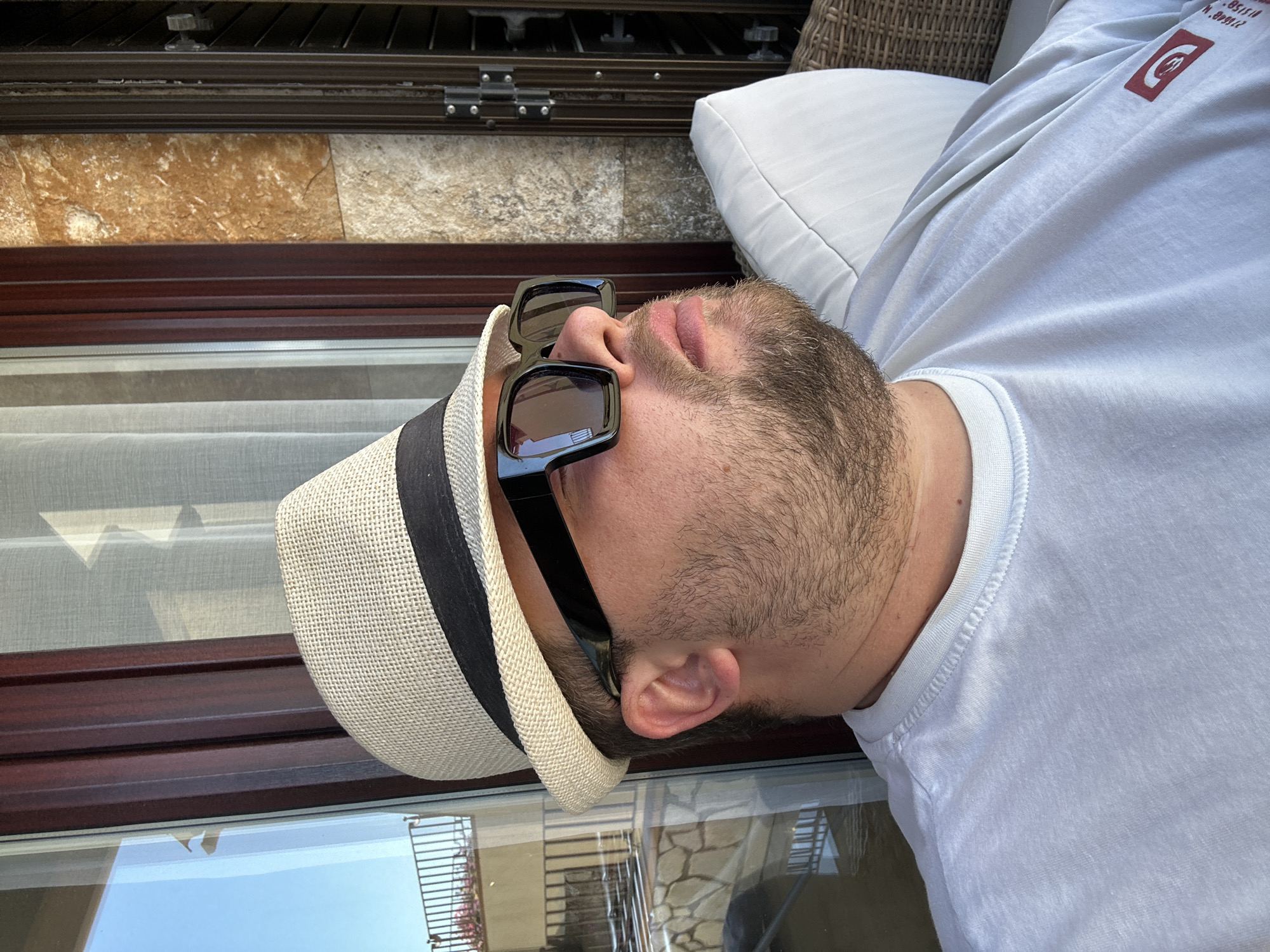 The image shows a man wearing sunglasses and a white straw hat with a black band, reclining on a white pillow. He is wearing a white t-shirt with a red logo on the shoulder. The background includes a window frame with a view outside, showing a balcony with a metal railing. The exterior features a textured stone wall. Based on the architectural details, particularly the stone wall, the location might be in Sant Feliu de Guíxols, Spain.