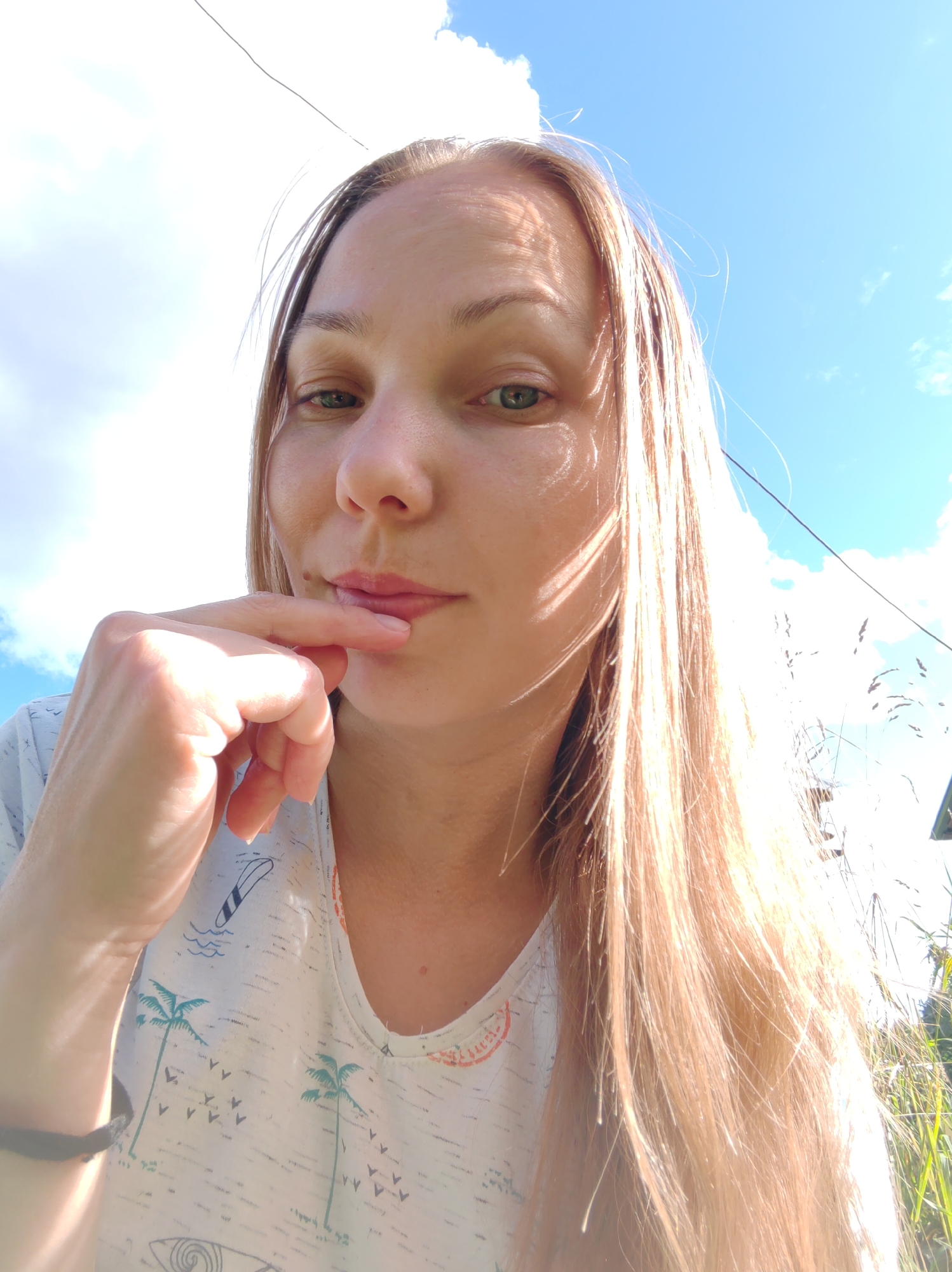 A close-up, selfie-style, outdoor photograph captures a woman in the city of Chukhlem, Russia. The woman, with long, blonde hair, is looking towards the camera with her right index finger touching her lips. She is wearing a white t-shirt adorned with a pattern of palm trees, surfboards, and small birds. The background consists of a bright blue sky with scattered white clouds, and hints of green grass and reeds suggest a natural, possibly rural or park-like setting within the city. A thin black wire stretches across the upper portion of the frame. The lighting is bright, indicating it is daytime.
