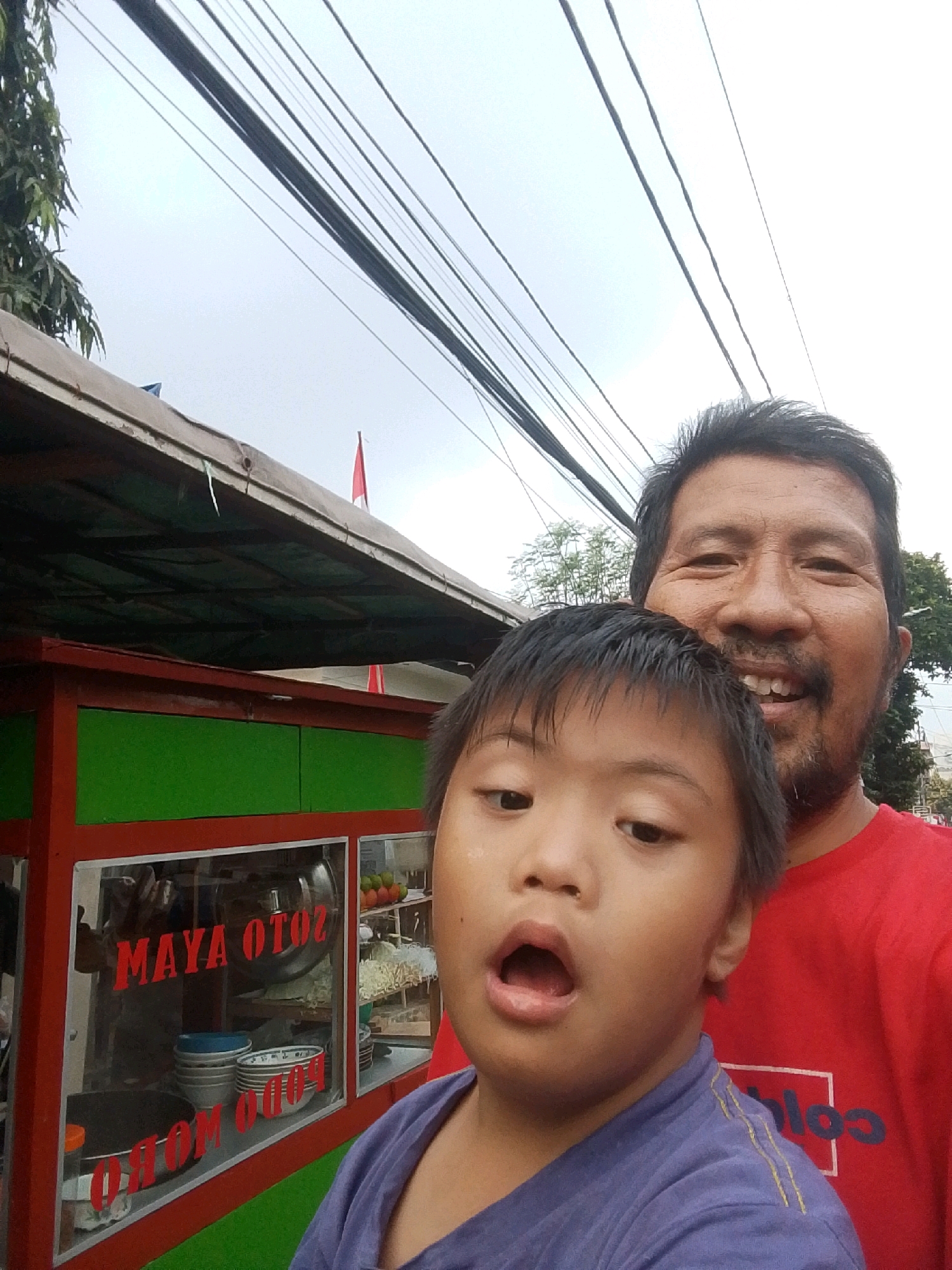 In a selfie taken outdoors, a man and a child stand in front of a food cart. The man, with dark hair and a beard, smiles broadly, his head positioned behind and slightly to the side of the child. The child, who appears to have Down syndrome, looks towards the camera with an open mouth, wearing a purple shirt. The food cart, painted red and green, has glass windows displaying bowls and food items. Red lettering on the window reads "MAYA OTOZ" and "OTOM ODOM". Overhead, a dense network of electrical wires crisscrosses the sky. In the background, a tree and buildings are visible, suggesting an urban environment. The overall scene conveys a casual, everyday moment, possibly in East Jakarta, Indonesia, given the presence of the Indonesian flag on the cart and the style of the food cart.