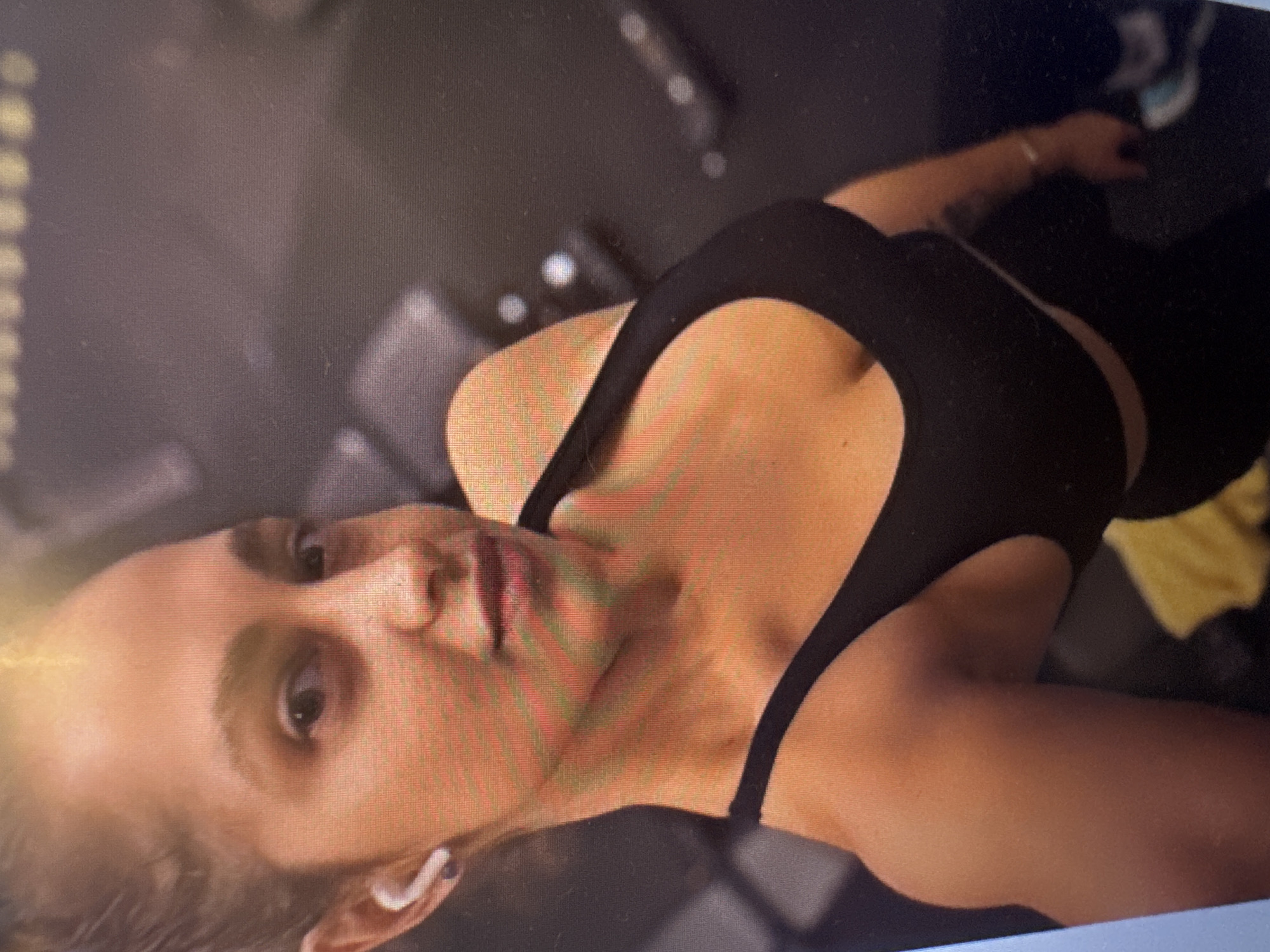 The image shows a close-up, slightly angled, selfie-style shot of a woman in athletic wear. She has her hair pulled back and is wearing white wireless earbuds. She is looking towards the camera with a neutral expression. Her attire consists of a black sports bra and black leggings. The background is dark and appears to be a gym setting, with some blurred equipment visible, including what looks like weight machines. There is no indication of the specific location being in the city of Murcia, Spain, or any other geographical markers.