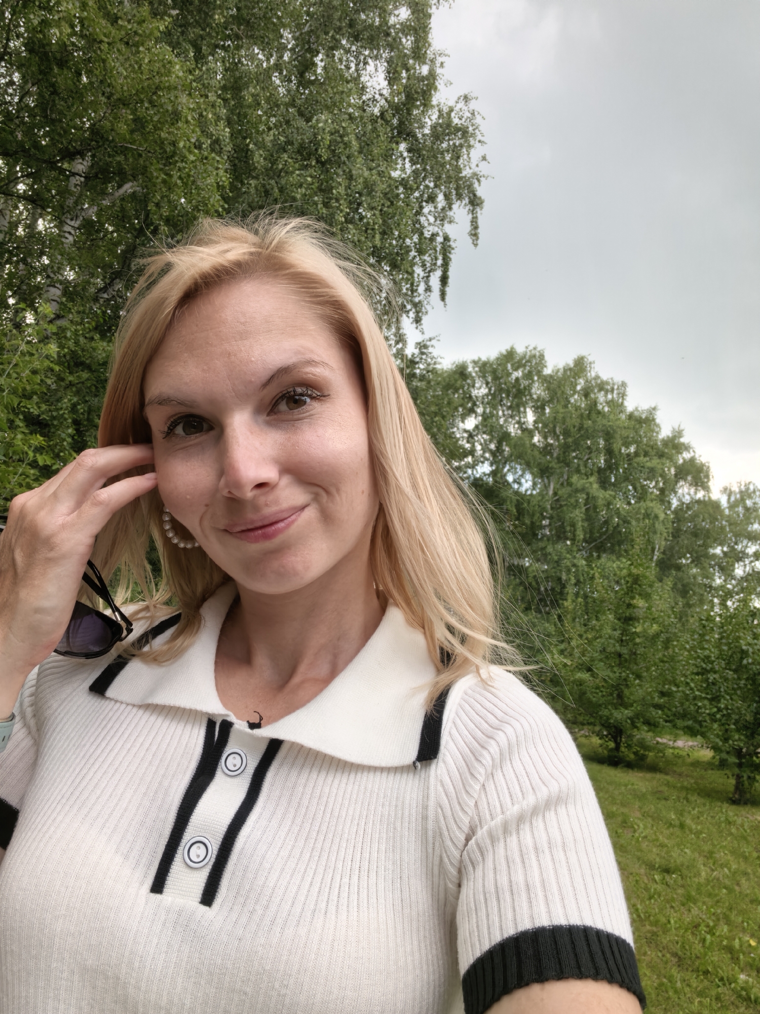 In a medium shot selfie, a blonde woman with fair skin and light brown eyes smiles at the camera. She is outdoors, with lush green trees and a cloudy sky in the background. She has her right hand up to her ear, and is wearing a white polo shirt with black trim and buttons. She also has on large pearl hoop earrings. The overall impression is that of a casual portrait taken in a park-like setting. The location is Sterlitamak, Russia.