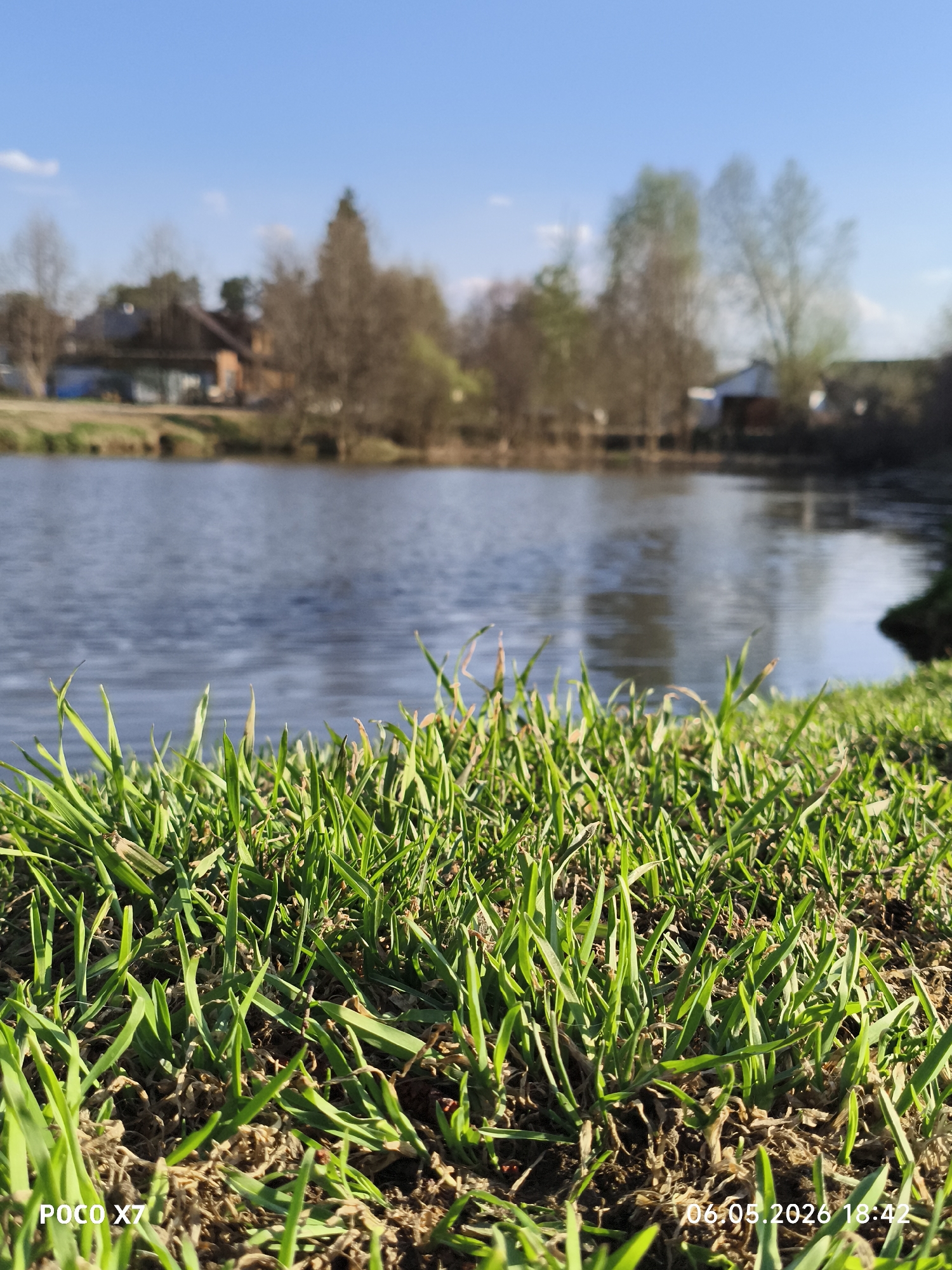 A close-up, eye-level shot depicts a grassy embankment near a body of water, likely a pond or lake. The foreground is dominated by vibrant green grass blades interspersed with brown soil. Beyond the grass, the dark blue water stretches across the midground, reflecting the daylight. In the blurred background, structures resembling houses and bare trees are visible. The sky is a clear, light blue. A watermark at the bottom left corner of the image reads "POCO X7," and the date and time "06.05.2026 18:42" are displayed in the bottom right corner. The location is Chaikovsky, Russia.