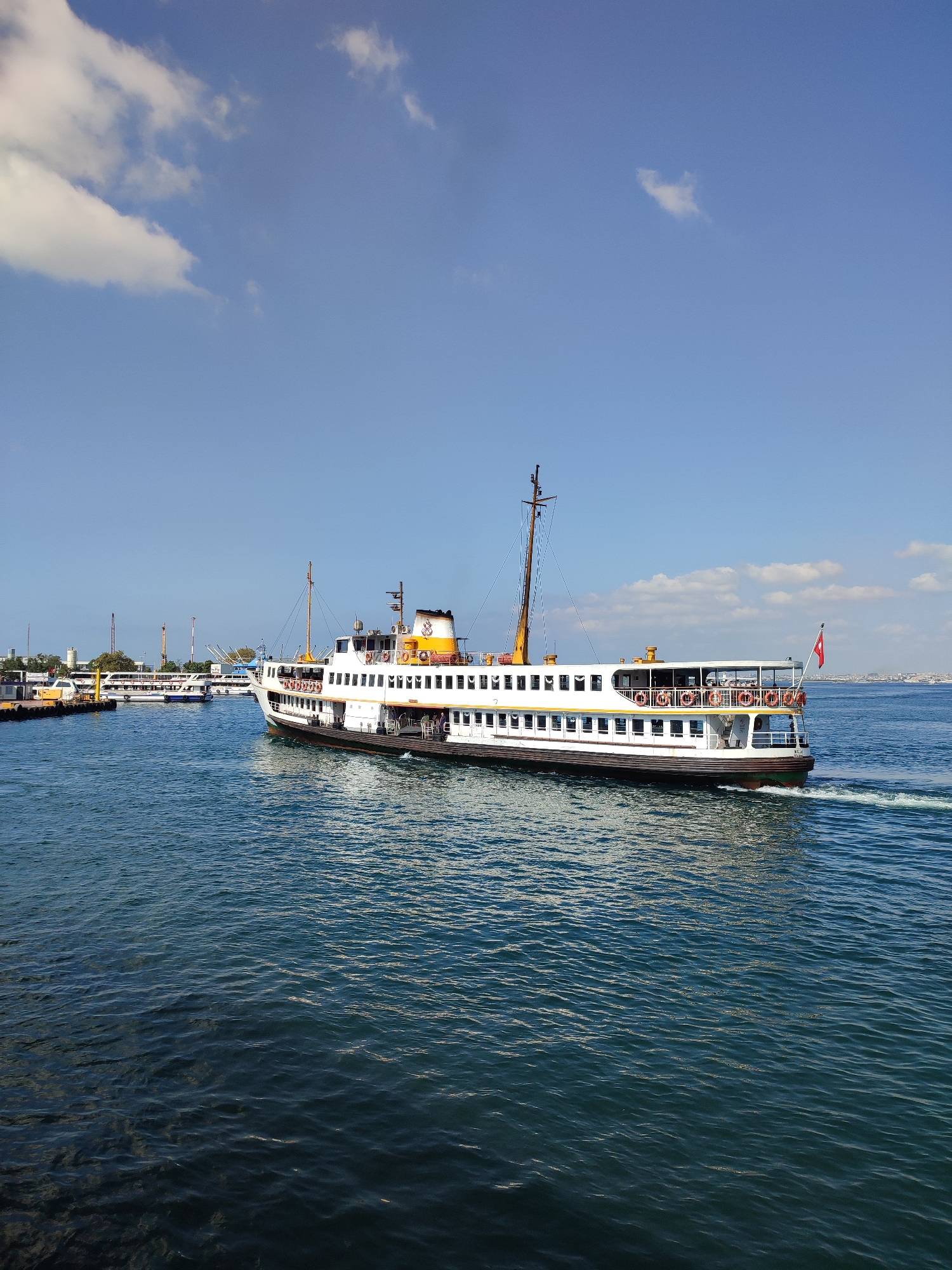 In the city of Kadıköy, Turkey, a passenger ferry navigates through the blue waters of the Bosphorus. The ferry, a large white vessel with multiple decks and windows, is adorned with orange life preservers and a Turkish flag at its stern. It is moving from left to right, creating a white wake in its path. In the background, other boats are visible, along with the distant cityscape. The sky above is a clear blue, dotted with fluffy white clouds. The water is choppy, reflecting the sunlight. The scene depicts a typical day of maritime transport in Istanbul's vibrant port.