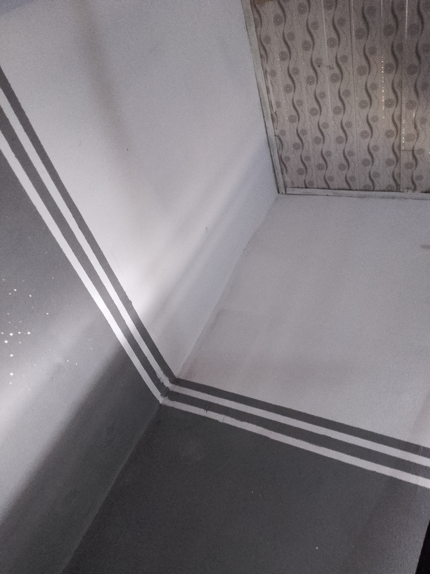 The image shows an interior corner of a room where two walls meet. The walls are painted with a two-tone design, with the lower portion being dark gray and the upper portion being white. A decorative stripe runs along the corner where the two walls meet, consisting of two parallel gray bands separated by a white stripe. The ceiling is visible in the upper part of the image and has a patterned design with wavy vertical lines and circular motifs. The lighting in the room casts shadows and highlights on the walls. There are no people or specific objects clearly identifiable in the scene. The overall impression is of a neutrally decorated interior space.