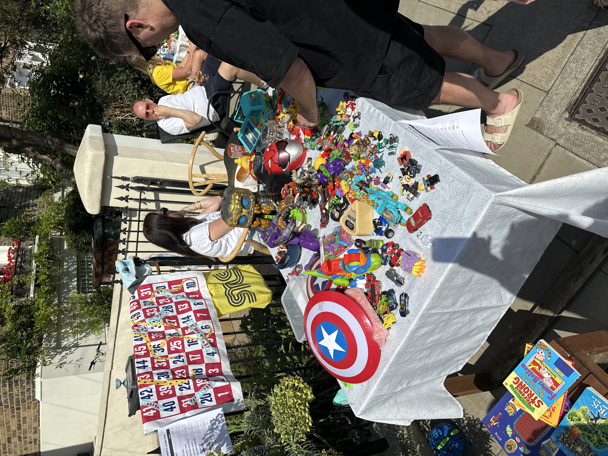 The image shows a table covered in a white tablecloth, displaying a collection of toys for sale at what appears to be a jumble sale. The toys include action figures, cars, and a Captain America shield. Beside the table, a game mat with numbered squares from 1 to 45 is placed, and some children's books are on the side. People are visible around the table, appearing to be browsing and examining the items on display. The location is likely in London, United Kingdom, based on visible signs, although further context would be needed to confirm the location with certainty.