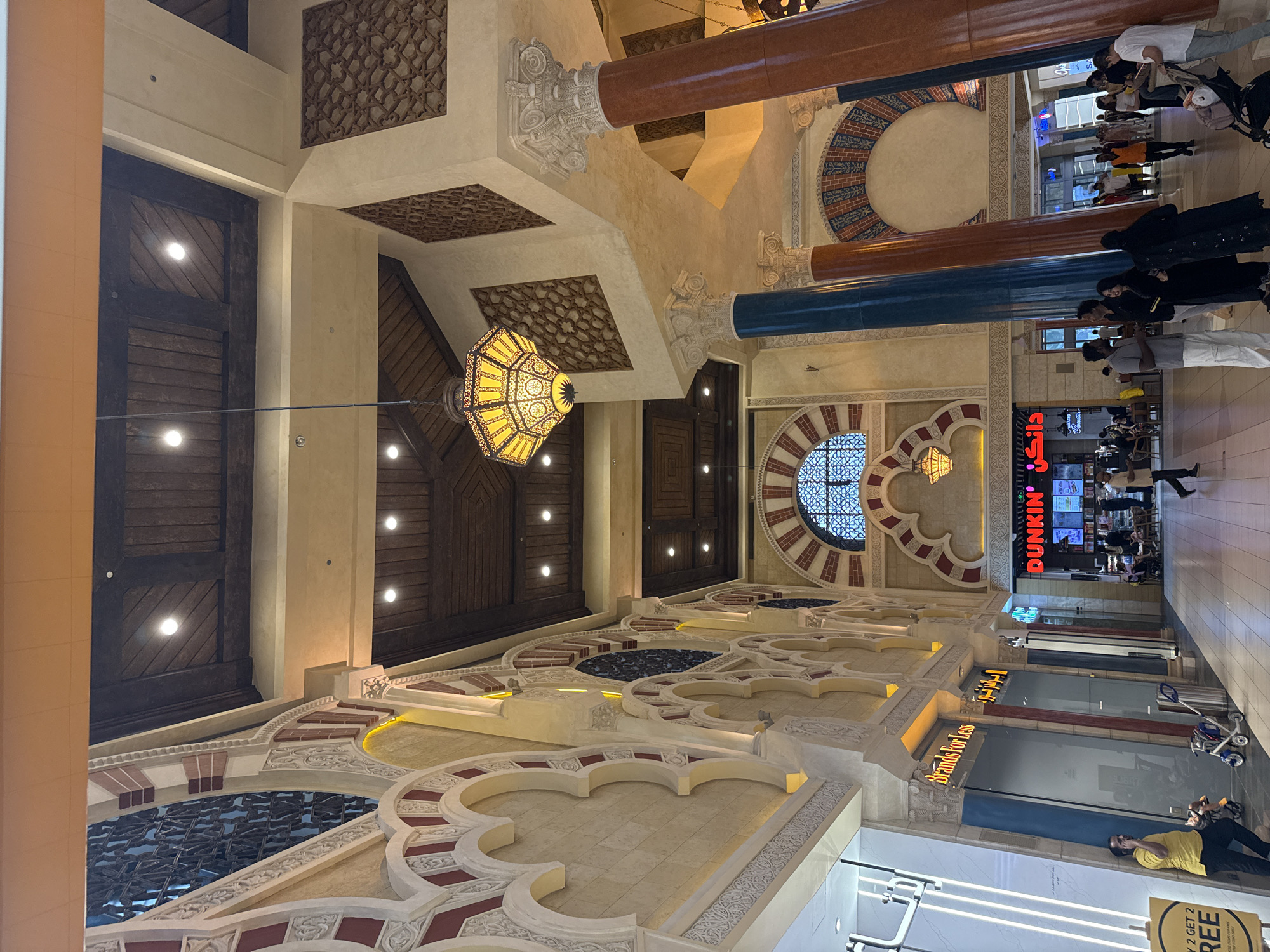 This image captures the interior of a shopping mall in Dubai, United Arab Emirates, characterized by its ornate, Moorish-inspired architecture. The foreground features a wide, tiled walkway with several people strolling and congregating. To the left, a woman in a yellow shirt stands near a sign offering a "Buy 2 Get 2 FREE" promotion, while a stroller is parked nearby. Further into the mall, several shops are visible, including "Brands For Less" and "Dunkin'". The mall's interior design is a prominent feature, with elaborate arched doorways, decorative latticework on the walls, and large, segmented columns in earthy tones. Suspended from the high, wood-paneled ceiling are intricate, lantern-style light fixtures. The overall impression is one of a grand and architecturally rich commercial space.
