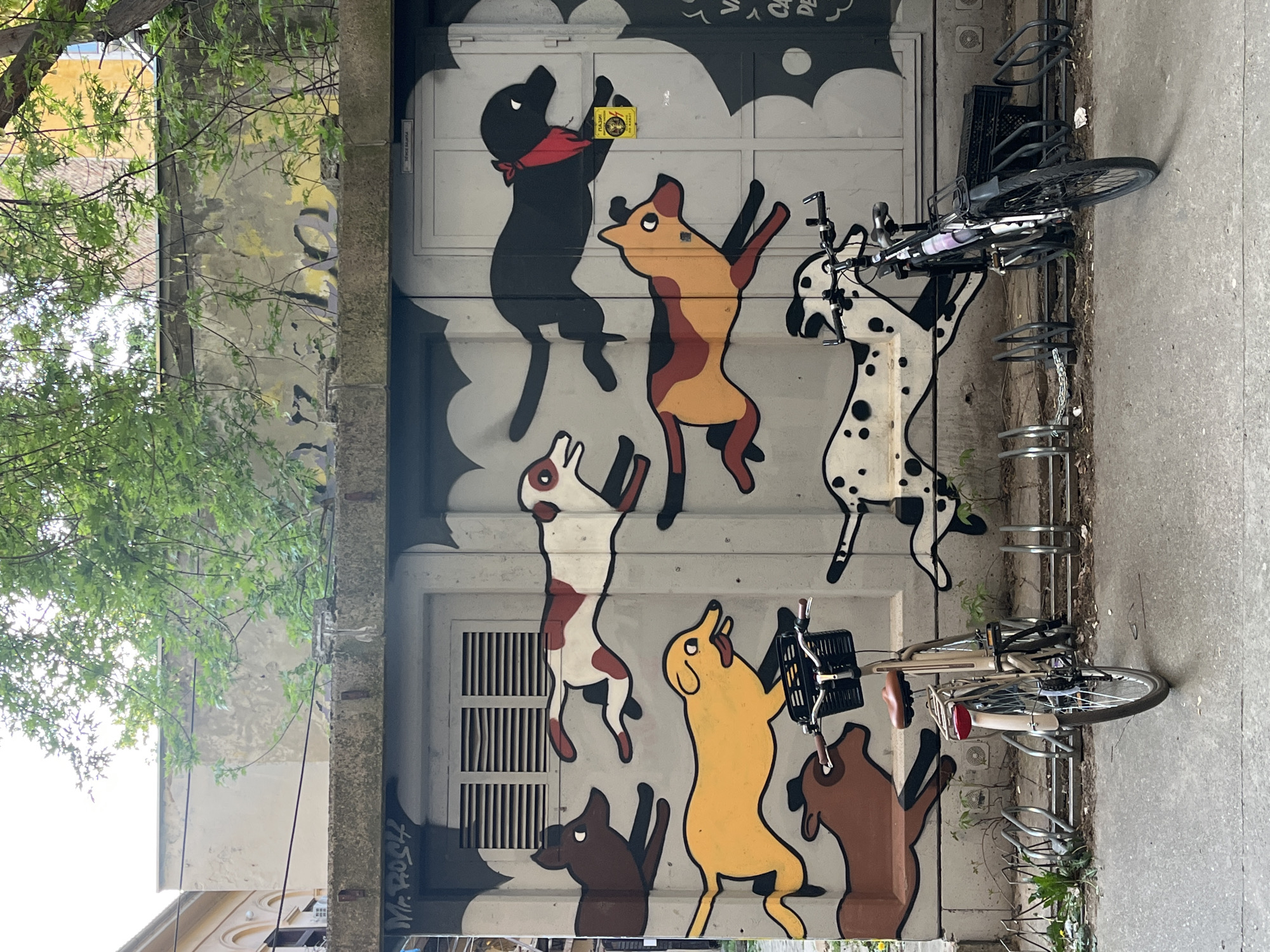 The image shows a mural painted on a building wall in Novi Sad, Serbia. The mural depicts several cartoonish dogs in various poses, seemingly dancing or jumping. One dog is black with a red bandana and appears to be holding a yellow object. Other dogs are brown, yellow, white with brown spots, and a Dalmatian. The background of the mural features cartoonish clouds.
A bicycle rack with two bicycles is positioned in front of the mural. The building exterior is slightly weathered with some graffiti. Trees and foliage partially obscure the left side of the mural and building. The scene is outdoors on a paved surface, likely a street or sidewalk.