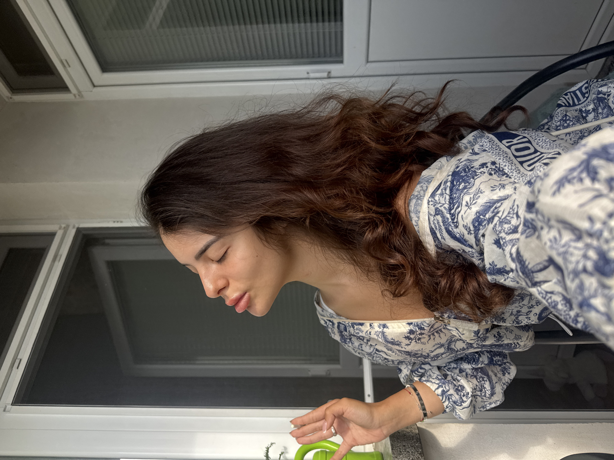 A young woman with long, wavy brown hair is on a balcony with white window frames and blinds. She is wearing a blue and white patterned top. Her eyes are closed and her lips are pursed. Her right hand is raised, with fingers slightly spread. A green watering can is visible to her left. The scene does not provide enough information to determine the city of Plovdiv, Bulgaria.