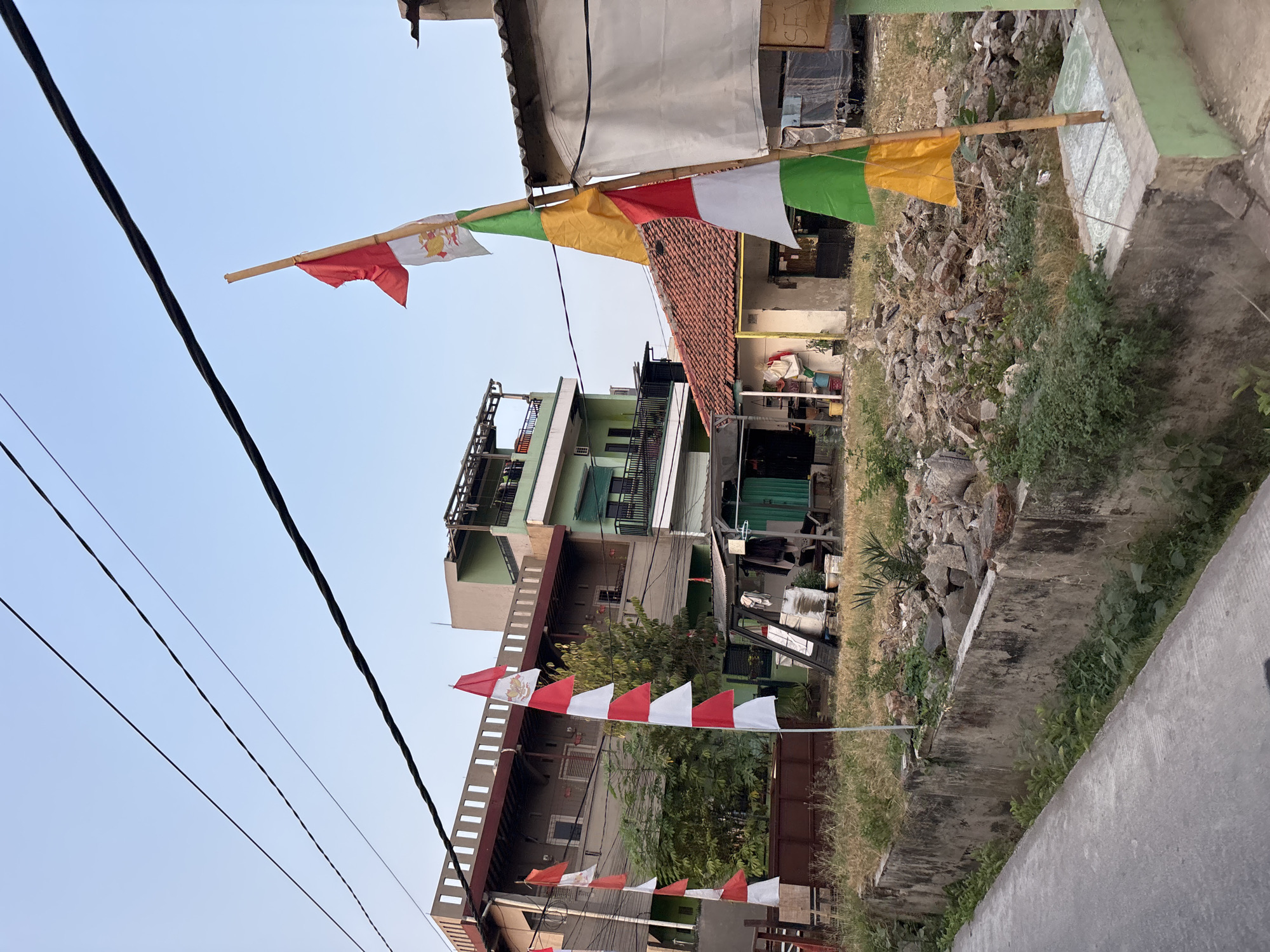 This is a full-shot, eye-level, outdoor vertical photograph of a street in Bekasi, Indonesia, featuring several Indonesian flags displayed on bamboo poles. The flags are red and white, representing the Indonesian national flag, and are arranged in a festive manner. Behind the flags, there are buildings, a narrow concrete drainage ditch, and a pile of rocks and debris. The sky is clear and blue. The overall scene appears to be a residential area, possibly decorated for a national holiday.