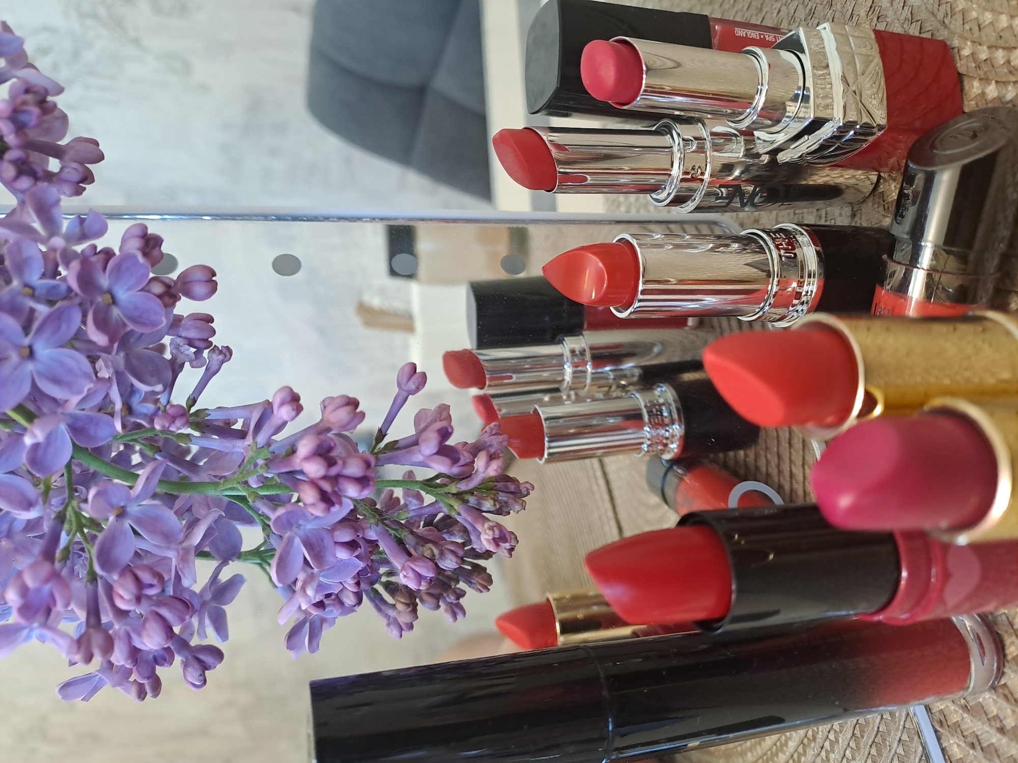 The image shows a still life arrangement of lipsticks and lilac flowers on a reflective surface. The lipsticks are various shades of red and pink, with different packaging designs. The lilac flowers are purple and arranged to the side of the lipsticks. The reflective surface creates multiple reflections of the objects, adding depth to the composition. The background is blurred and indistinct. There is no indication of Bel'kovo, Russia.