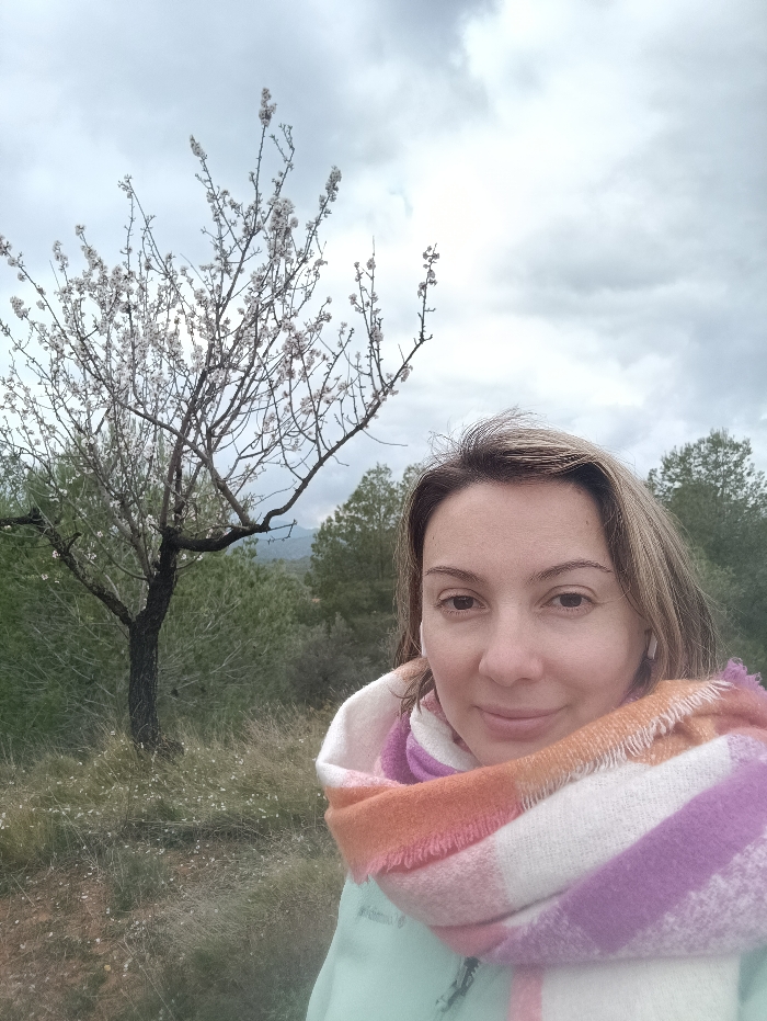 A female subject is depicted in a close-up, outdoor portrait in Jérica, Spain. She has light brown, shoulder-length hair and is looking directly at the camera with a subtle smile. She is wearing a light mint-green jacket and a large plaid scarf with white, orange, pink, and purple stripes wrapped around her neck and shoulders. Small white earbuds are visible in both ears. Behind her, positioned to the left, stands a flowering tree with white blossoms, likely an almond tree, rooted in dry grass with scattered small white flowers. The mid-ground and background consist of dense green foliage, primarily evergreen trees, and distant hills under a heavily overcast, cloudy sky.