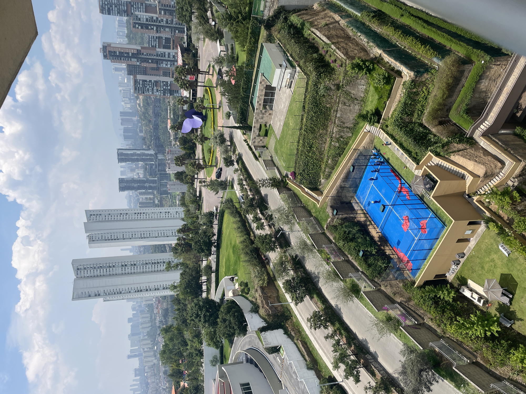From an elevated vantage point in Huixquilucan de Degollado, Mexico, the image captures a dense urban landscape juxtaposed with pockets of manicured greenery. Numerous high-rise apartment buildings dominate the skyline in the distance, shrouded in a slight haze. Closer to the foreground, the image reveals a meticulously planned environment with paved walkways lined with trees and manicured lawns. A blue paddle tennis court with red markings is visible, adjacent to a building and open grassy areas. Terraced landscaping ascends, creating a visually layered effect. An oversized purple art installation stands near a road with sparse traffic. The scene suggests a controlled, upscale environment within the broader urban context.