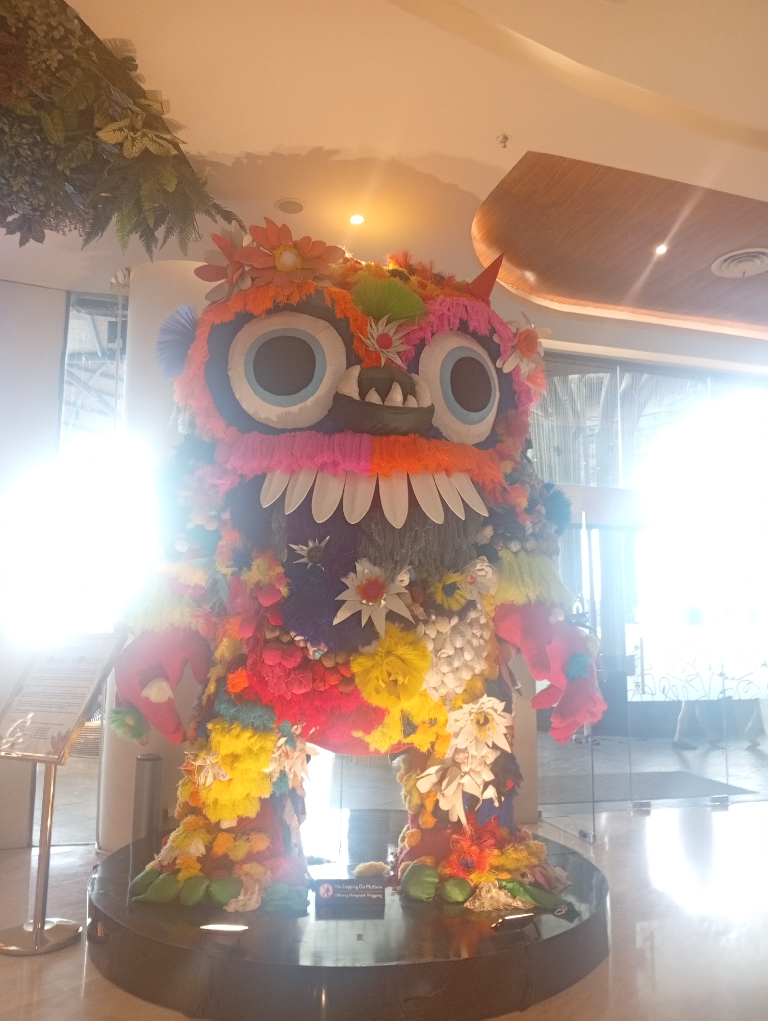 The image shows a colorful monster sculpture in the lobby of a building. The sculpture has large eyes, sharp teeth, and is covered in flowers and other materials of various colors. It stands on a black circular platform with a sign that says "No Stepping On Platform Dilarang Menginjak Panggung". The lobby has a modern design with marble floors, glass walls, and an artificial plant hanging from the ceiling. The location is Denpasar, Indonesia.